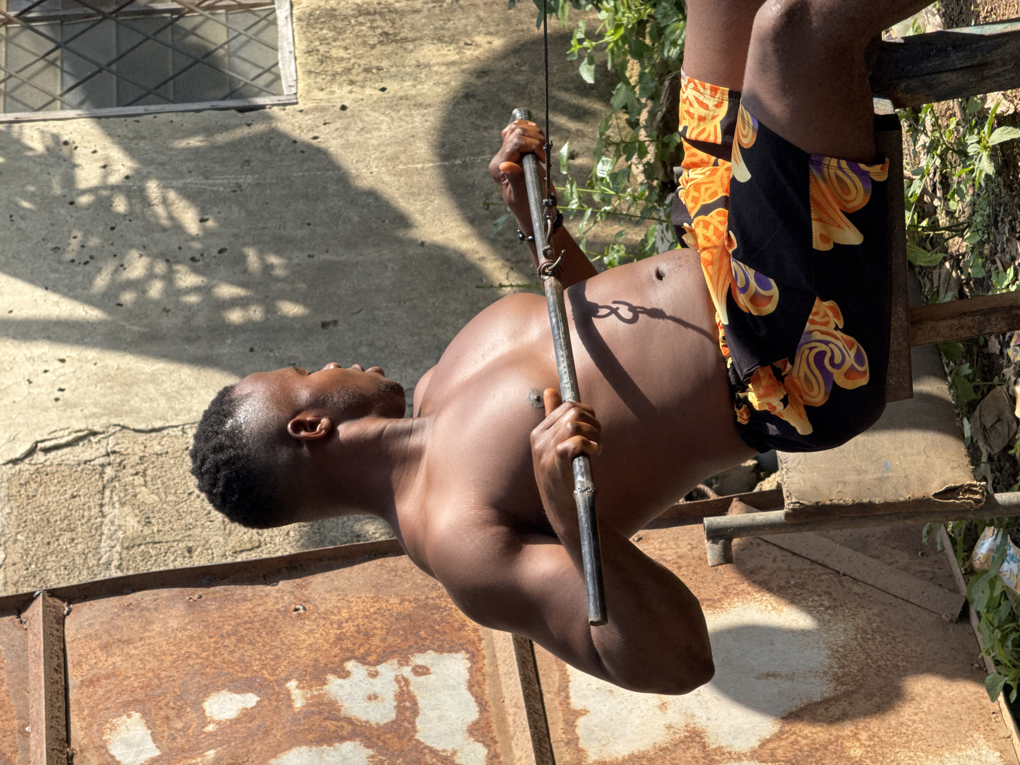 A shirtless Black man is performing a rowing exercise outdoors in Akwanga, Nigeria. He is seated on a metal stool, holding a metal bar attached to a cable. He is wearing black shorts with an orange and purple patterned print. Behind him, a textured concrete wall is visible, with shadows cast by foliage on its surface, and a window with a metal grate. To his left, a large, rusty metal surface dominates the background. Lush green plants and some debris are present on the ground around the stool. The lighting suggests it is daytime, with strong sunlight creating distinct shadows.