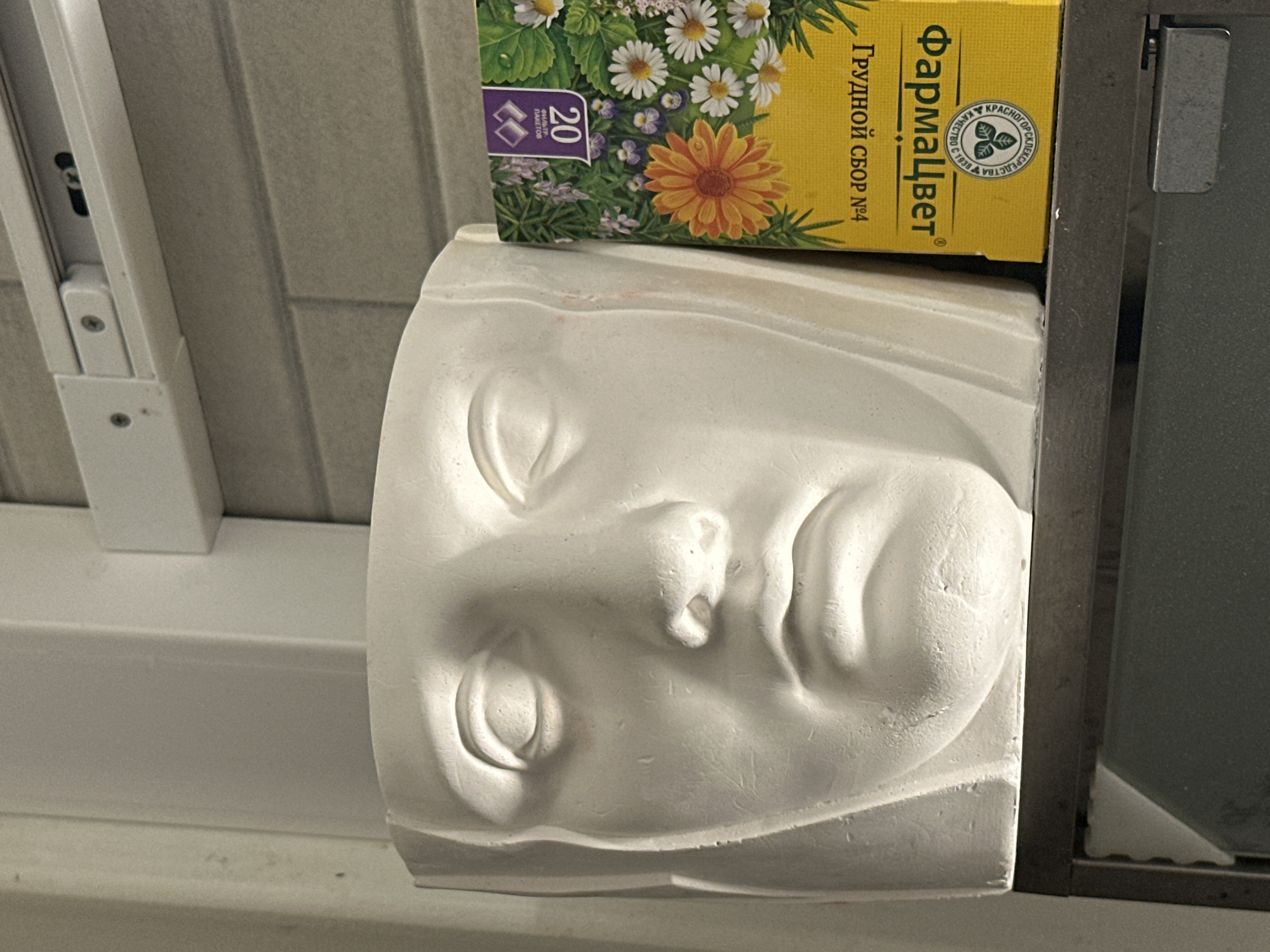 A still-life image presents a white, plaster sculpture of a face lying on its side, with closed eyes and prominent facial features. Resting on top of the sculpture is a rectangular yellow box of "Фармацвет" herbal tea, adorned with floral patterns and Russian text indicating its contents. The objects are positioned near a window frame, suggesting an indoor setting. The location, Новоглаголево, Russia, is not visually discernible from the objects and scene depicted in the image.