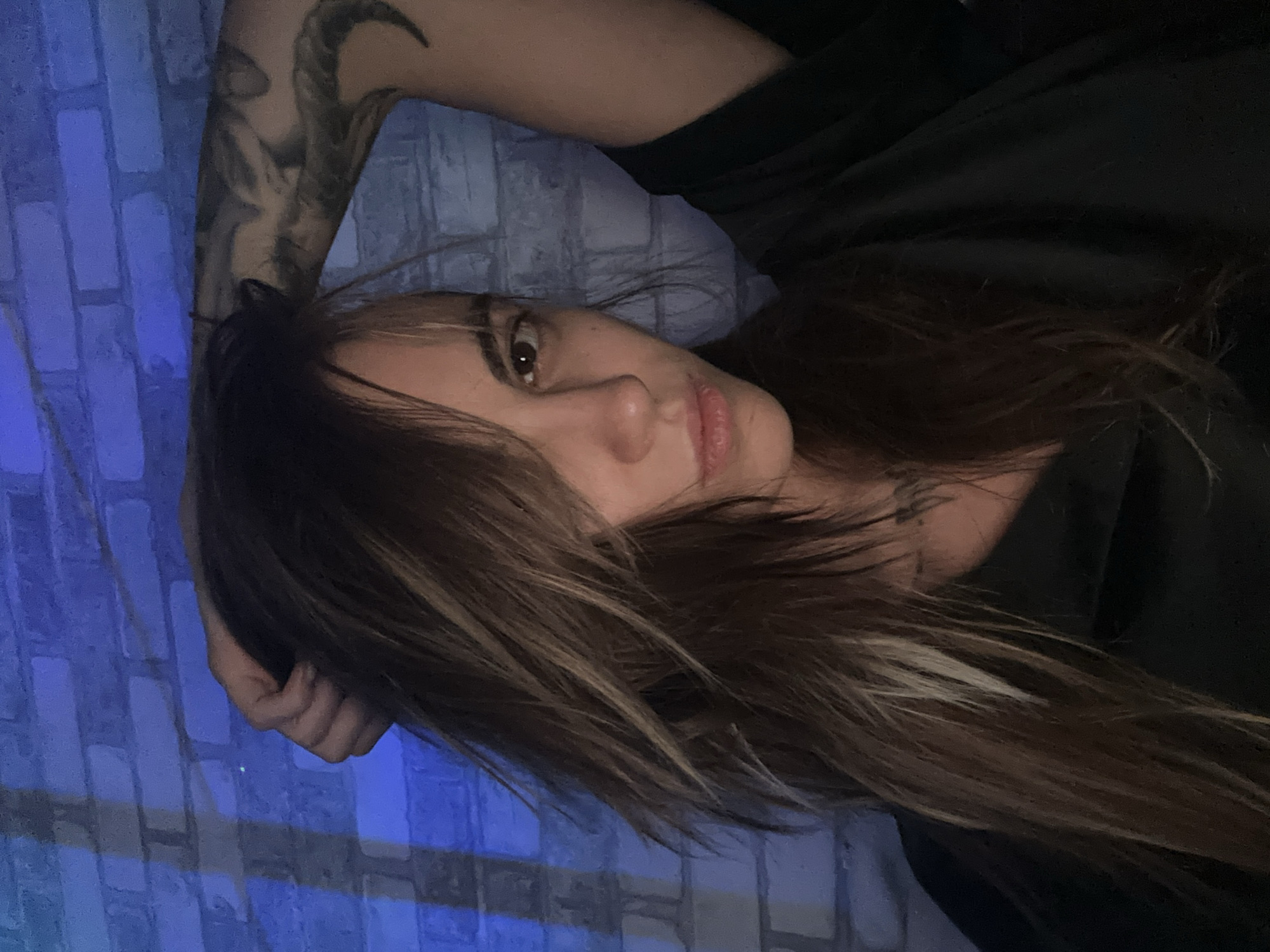 A woman is captured in a close-up shot, her face angled towards the camera. She has long, layered brown hair with lighter streaks, and her left hand is raised, resting on her head. Her left arm is visible, adorned with a dark tattoo. She is wearing a black top. The background consists of a light-colored brick wall illuminated by blue light, casting shadows. The overall scene appears to be indoors. The location in Moscow, Russia is not discernible from the provided image.