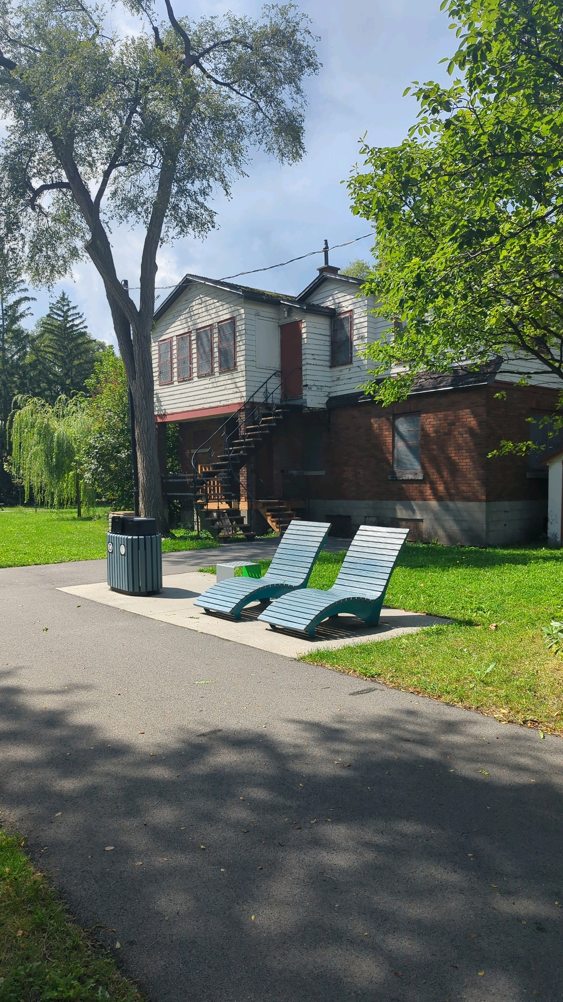 The image depicts an outdoor scene with a building, lounge chairs, a trash receptacle, a pathway, and a grassy area. The building appears to be a two-story structure with white siding on the upper portion and red brick on the lower portion. It has a set of external stairs leading to the upper level. Two light blue lounge chairs are positioned on a paved area in front of the building. A dark blue trash receptacle is located to the left of the lounge chairs. A paved pathway runs in the foreground, with a grassy area bordering it and extending towards the building. Trees are visible in the background, one large tree to the left and foliage on the right. The sky is partly cloudy. There are no people or animals visible in the image. The scene suggests a public park or recreational area.
