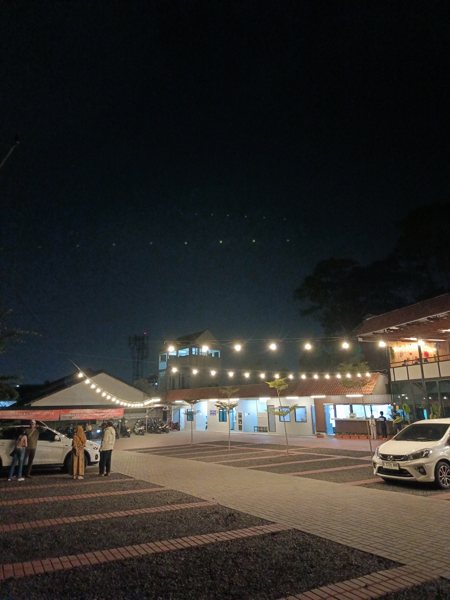 This is a nighttime, medium-shot, vertical photograph of a paved outdoor area in front of buildings. The scene appears to be a public space, possibly a food court or a gathering area, in Cimahi, Indonesia. In the foreground, a white car is parked on the right side of the frame, facing left. The car has license plate "D 1226 AHK". To the left of the car, there are three people standing near a white SUV. One person, wearing a yellow headscarf and yellow clothing, stands in front of the SUV. To their left, a person in a green shirt and dark pants is partially visible. To the right of the person in the yellow headscarf, another person in a light-colored jacket and dark pants stands.
The ground is paved with textured tiles and dark gravel. In the middle ground, a long, single-story building with an orange-tiled roof runs horizontally across the frame. String lights with warm yellow bulbs are hung across the area, illuminating the scene. Behind this building, a taller building with multiple windows is visible, also lit from within. Further back, a telecommunications tower can be seen against the dark night sky. The sky is dark blue and speckled with a few faint, out-of-focus lights. Trees are visible on the right side of the frame, silhouetted against the sky and buildings. The overall atmosphere is calm and inviting, with the lights creating a warm ambiance.