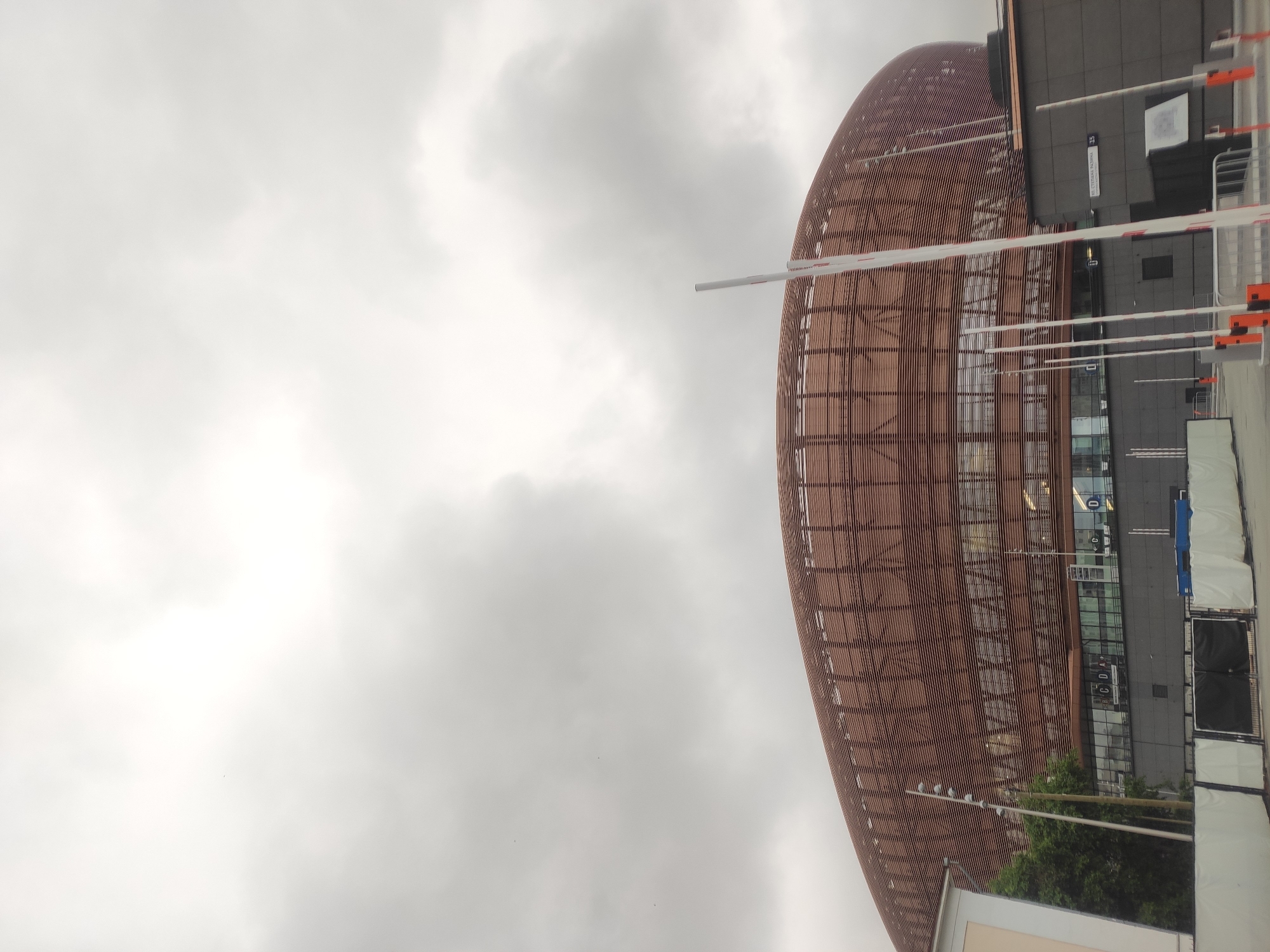 The image shows the exterior of a modern stadium in Yekaterinburg, Russia, under a cloudy sky. The stadium features a distinctive, curved facade composed of horizontal, reddish-brown louvers, creating a layered appearance. Large glass windows are visible behind the louvers on the lower levels. In the foreground, there are several white barriers with red accents, suggesting a controlled access area or event setup. A dark gray building with signage, including "ул. СТЕПАНА РАЗИНА 15" (Stepan Razin Street 15), is situated in front of the stadium. A single tree stands to the left of the stadium's entrance, partially obscured by a light-colored building. The overall impression is of a large, contemporary sports or entertainment venue.