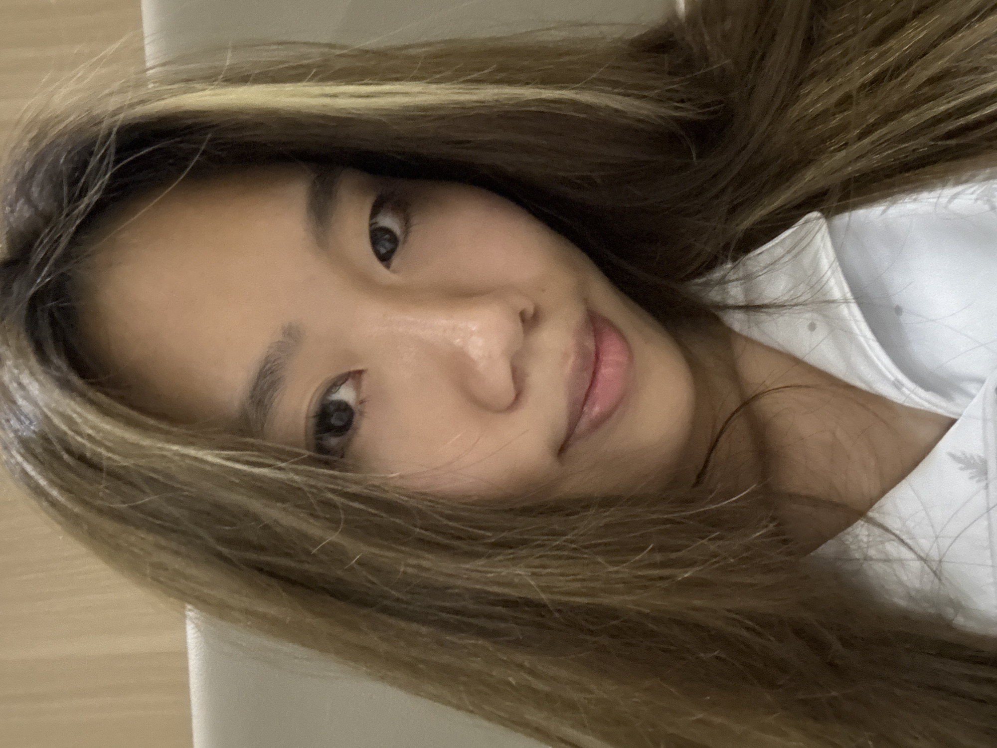The image is a close-up portrait of a young woman, likely taken indoors in Dunhuang City, China. She has long, flowing hair with shades of light brown and blonde highlights, which partially covers her forehead and spreads around her shoulders. Her head is tilted, and she gazes directly at the camera with a calm, gentle expression and a subtle, soft smile. Her lips have a natural pink hue, and her eyes are dark and open.
She appears to be wearing a white collared shirt or top, visible on the right side of the frame, which has a subtle textured or patterned detail. The background is simple and light-colored, suggesting an uncluttered indoor setting such as a room or bed area. The image provides no specific visual cues to determine the time of day or weather conditions, and no visible text is present. The overall mood conveyed is relaxed and peaceful.