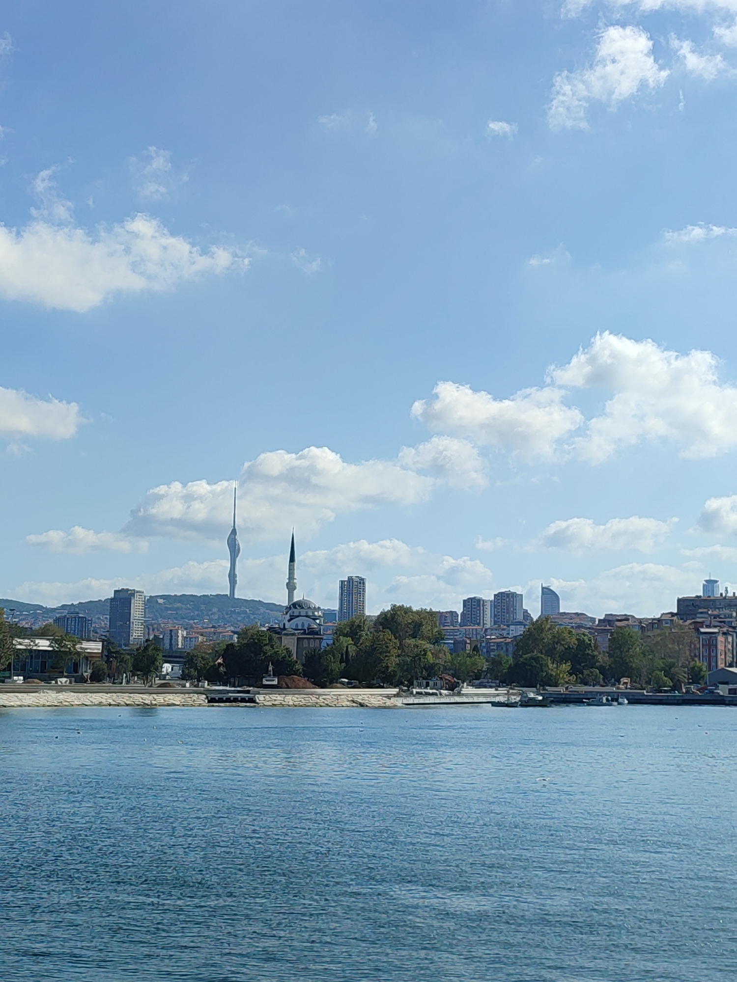 This is a landscape photograph of a city skyline across a body of water. In the foreground, a large expanse of calm, blue water occupies the bottom half of the image. Small ripples are visible on the surface. Along the far edge of the water, a paved embankment is present, with a line of trees and buildings beyond.
The skyline is diverse, featuring a mix of modern high-rise buildings and older structures. A prominent feature is a tall, slender tower with a distinctive curved top, resembling an antenna or observation tower, situated on a distant hill. To the right of this tower, a white mosque with a tall, thin minaret stands out against the sky. Several other taller buildings, appearing to be office buildings or residential complexes, are scattered throughout the skyline.
The sky is a clear, pale blue with scattered white clouds, some of which are puffy and well-defined, while others are wispy. The lighting suggests it is daytime, likely mid-morning or afternoon. The overall scene depicts an urban environment adjacent to a body of water, likely a bay or strait. This view is consistent with the coastline of Kadıköy, Turkey, which overlooks the Bosphorus strait and offers views of the Istanbul skyline. No people are clearly discernible in the image.