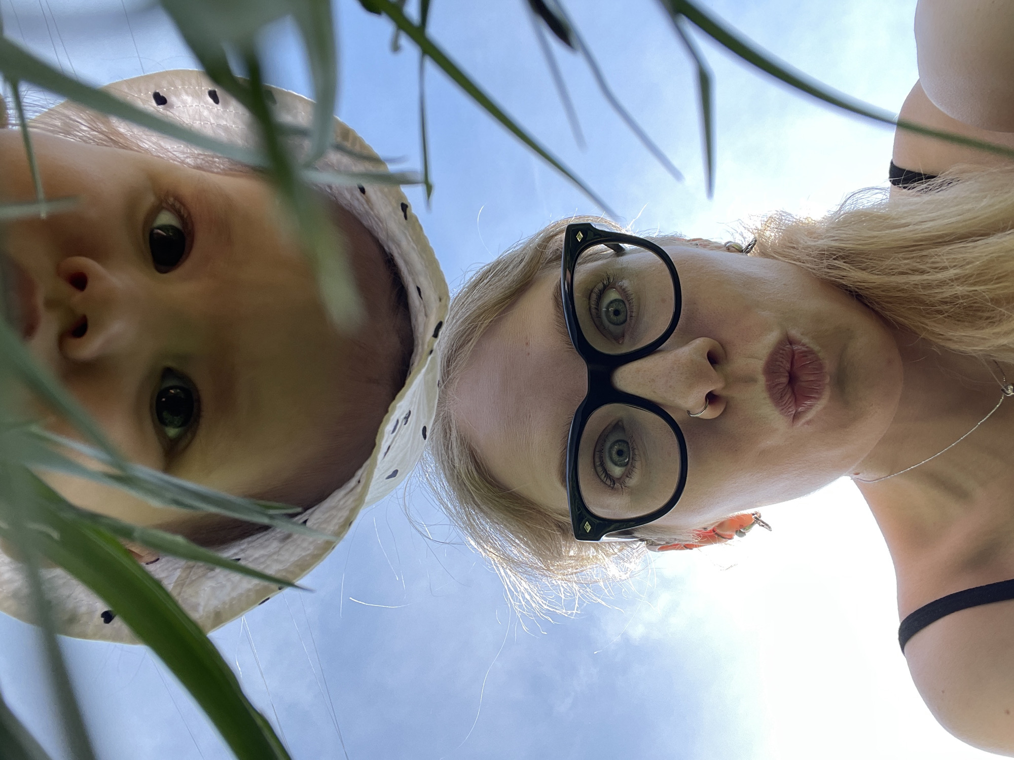 A low-angle shot depicts two individuals, a baby and an adult woman, presumably looking down at the camera. The baby is on the left wearing a white hat with black polka dots. The woman, on the right, is wearing black-framed glasses and has blonde hair. The background is a bright blue sky with light clouds, partially obscured by green foliage. A thin silver necklace is visible around the woman's neck. There is a black strap on her shoulder. The location is not discernible from the image and cannot be identified as Izhevsk, Russia.