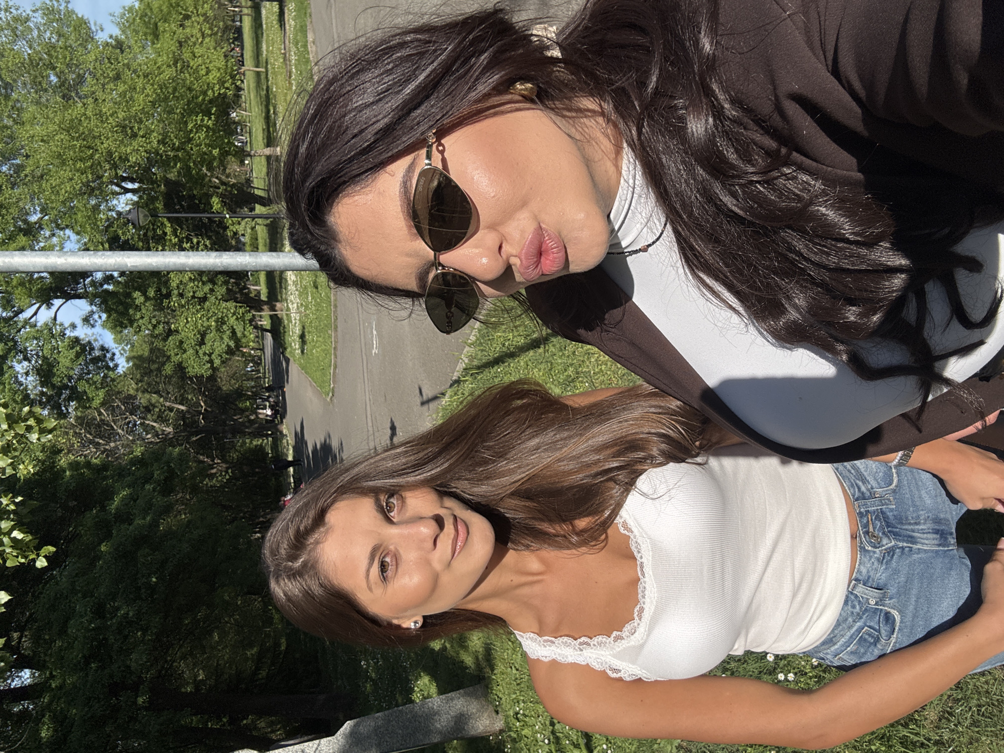 Two young women are in a selfie. The woman in the lower half of the image has light brown hair, is wearing a white lace-trimmed tank top, and jeans. The woman in the upper half of the image has long dark hair and is wearing sunglasses and a dark-colored jacket. The background appears to be a park with grass, trees, and a paved pathway. The location is not identifiable as Burgas, Bulgaria, based on the visual information provided.