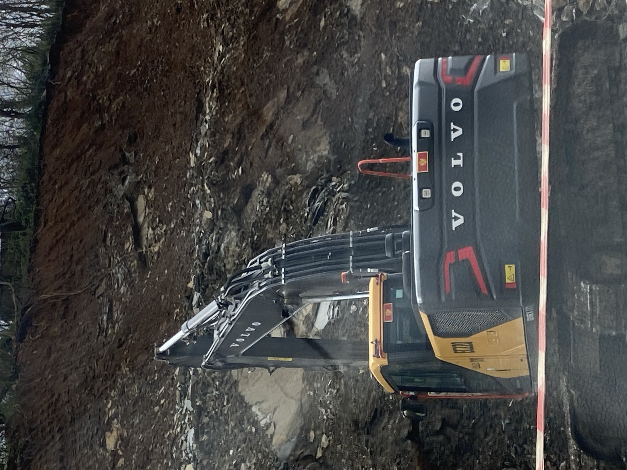 The image depicts a Volvo EC220 tracked excavator positioned at the base of a steep, excavated earth and rock embankment in Lastva Grbaljska, Montenegro. The excavator is dark grey with a yellow cab and lower side panel, featuring "VOLVO" branding on its boom and rear, and "EC220" on the cab side. A small Montenegrin flag emblem is visible on the right side of the excavator's cab roof. The machine appears stationary, with its boom extended upwards towards the slope. The embankment is composed of exposed dark soil, rock fragments, and stratified rock layers, rising sharply into the background. Sparse tree roots and bare branches are visible along the crest of the embankment. A red and white striped barrier tape runs horizontally across the foreground, positioned in front of the excavator's tracks, indicating a demarcated work zone. No individuals are present in the scene.