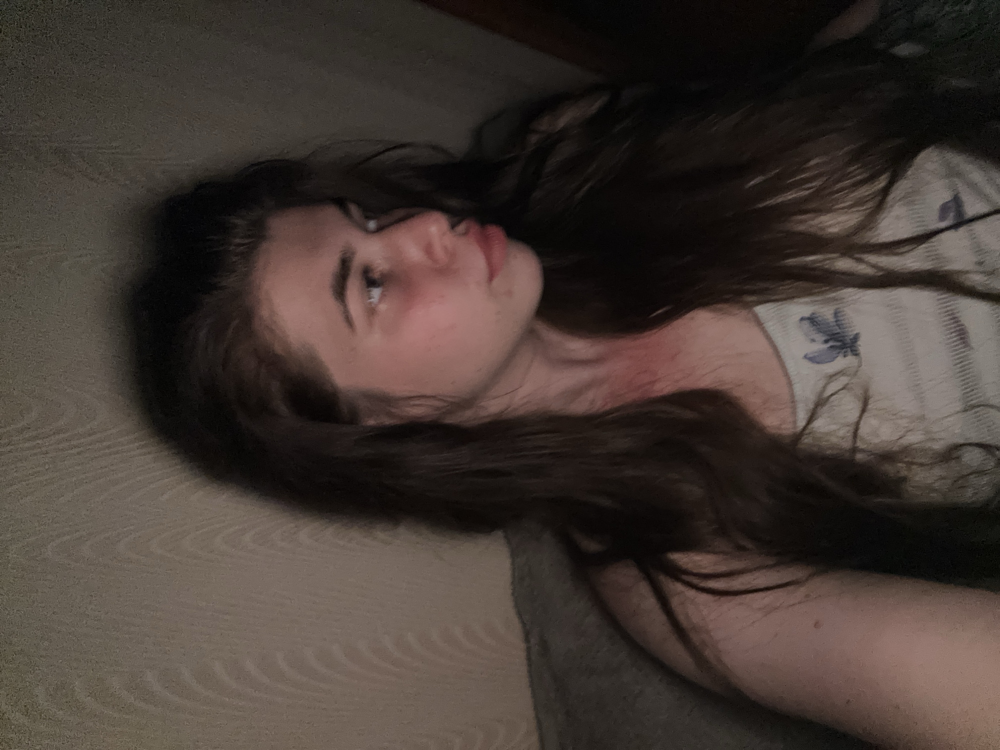 A young person with long, dark hair is shown in a close-up shot, looking away from the camera. They are wearing a light-colored tank top with a subtle pattern. The lighting is dim, with a reddish hue casting on their face, particularly on their cheek and neck area. The background appears to be a wall with a textured pattern. There is a dark object, possibly a piece of furniture or clothing, in the lower left corner of the image. The image quality is somewhat grainy, suggesting it was taken with a low-light setting or a lower-resolution camera. The scene does not contain any explicit geographical markers that would indicate its location in the city of Мещёры, Russia.