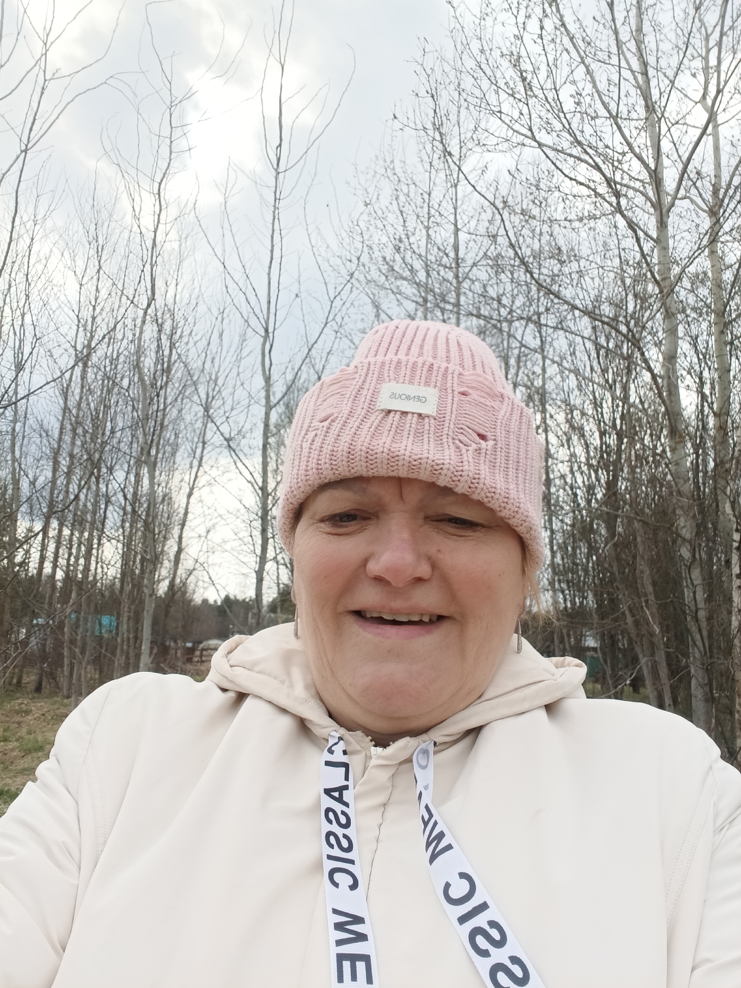 A woman in Красный Маяк, Russia, is taking a selfie outdoors. She is wearing a pink knitted beanie with a white tag, and a light beige hooded jacket with a white drawstring that has the words "CLASSIC ME" printed on it. The background consists of a wooded area with thin, bare trees against a cloudy sky. There's a hint of green grass visible at the bottom of the frame.