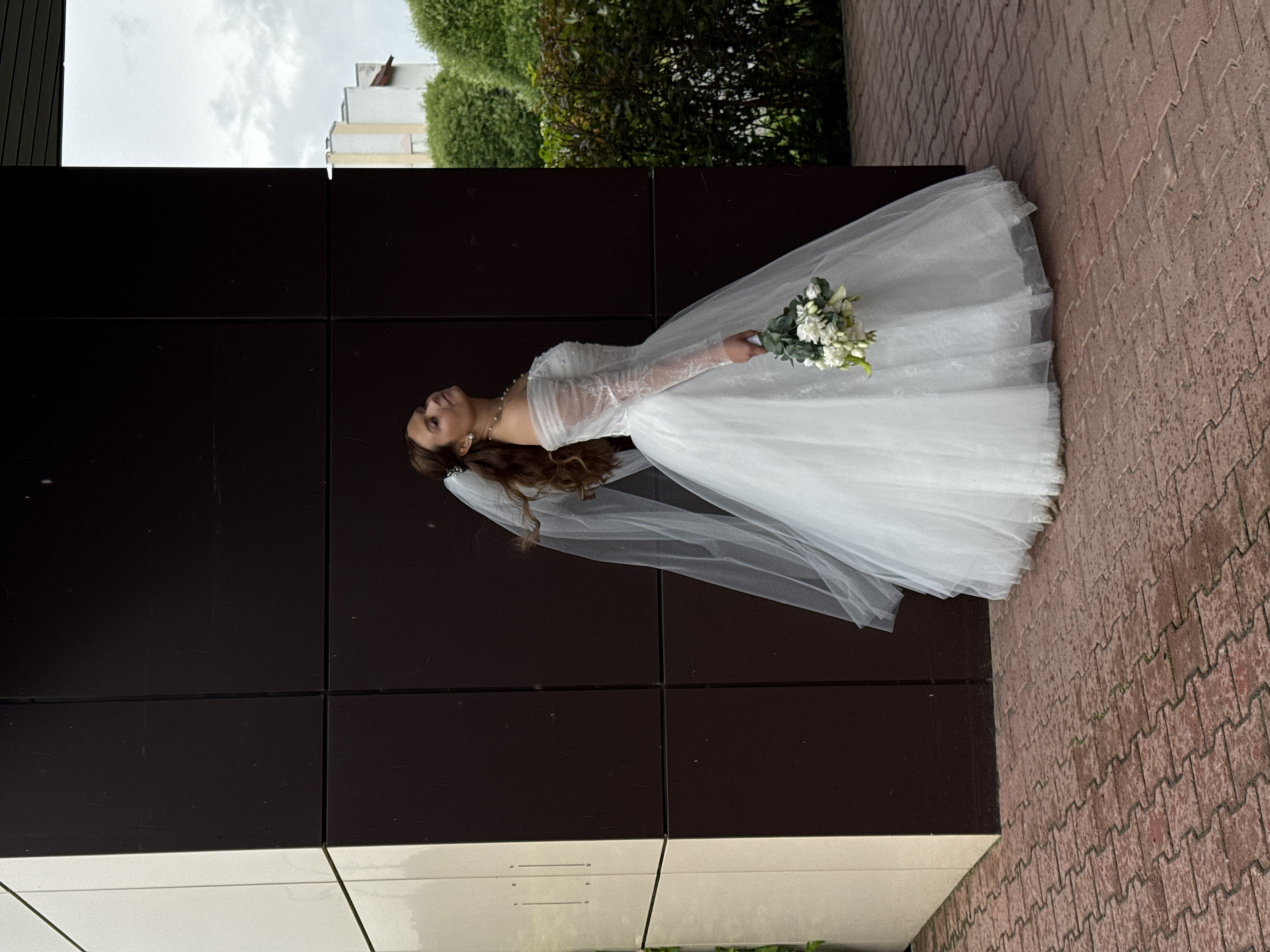 A full-length outdoor portrait captures a bride posing in Chaikovsky, Russia. The central subject is a woman with long, flowing brown hair, adorned with a white veil that extends down her back. She wears an elegant white wedding gown featuring long, sheer sleeves with delicate embroidery, and a simple pearl necklace. Her gaze is directed upwards, and she holds a bouquet of white flowers and green foliage in her left hand, which rests gently at her side.
The woman stands upright against a dark brown, multi-paneled building facade, which occupies a significant portion of the background. To the right of the bride, the ground is covered with reddish-brown pavers arranged in a herringbone pattern. In the upper left background, above the dark building, an overcast sky is visible, along with glimpses of a white building and lush green bushes. The lighting is soft and diffused, indicating an overcast day. The overall scene suggests a posed wedding photoshoot. No text is visible in the image.