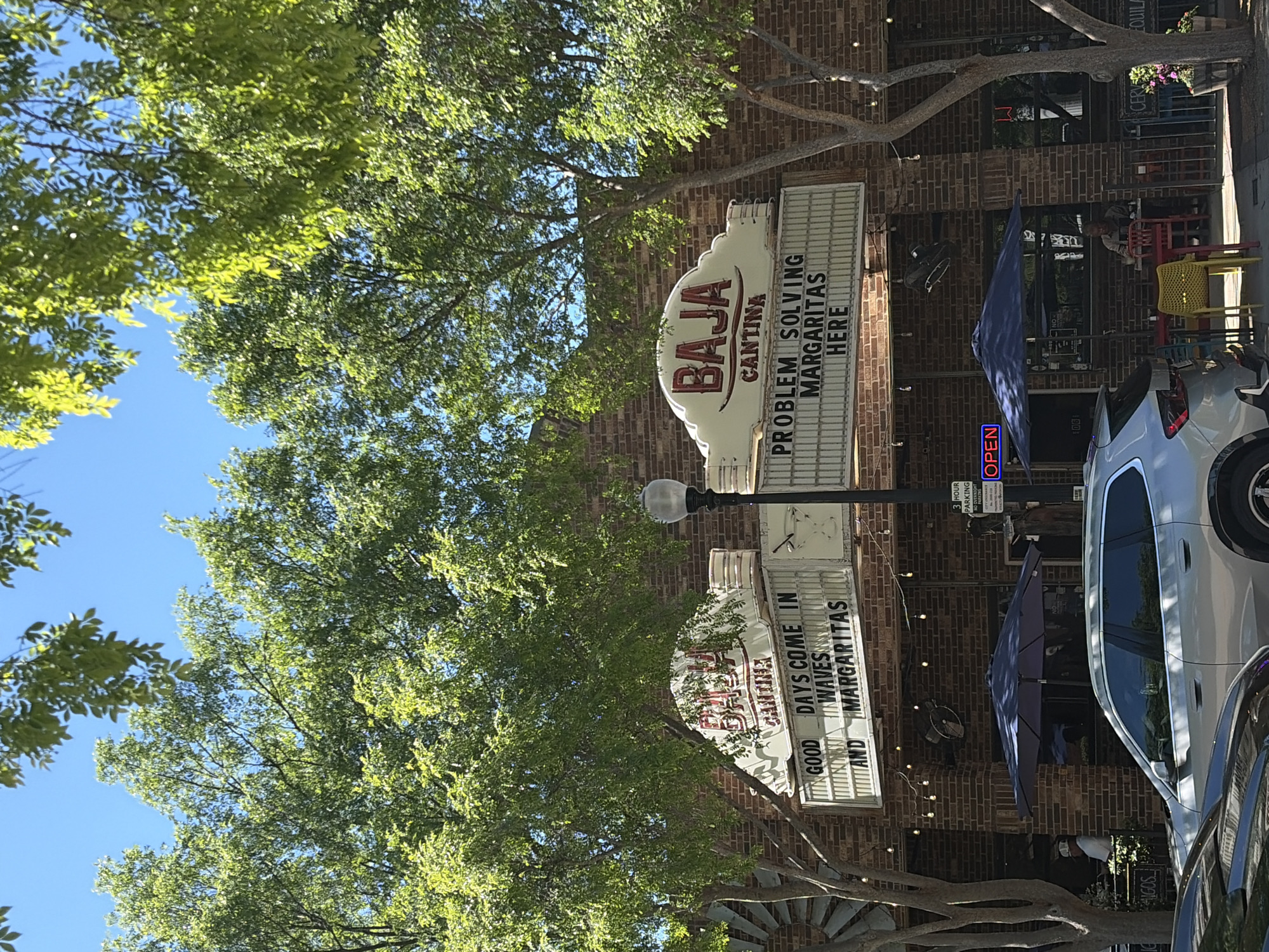 The image depicts the facade of "Baja Cantina", a restaurant in what appears to be an urban area. The building has a brick exterior, with two vintage-style lighted signs. The upper sign reads "Baja Cantina: Problem Solving Margaritas Here", and the lower sign reads "Good Days Come In Waves And Margaritas."
An "Open" sign is displayed near the restaurant's entrance, which is shaded by a partially opened blue umbrella. A silver car is parked along the sidewalk, partially obstructing the view of the restaurant's outdoor seating. Trees with full green foliage are positioned on either side of the building, partly obscuring the view.
The image was taken during daylight hours, as indicated by the bright blue sky and clear lighting. There is a 3-Hour Parking sign next to the entrance, indicating this might be located in a busy area. The location is Roanoke, United States of America.