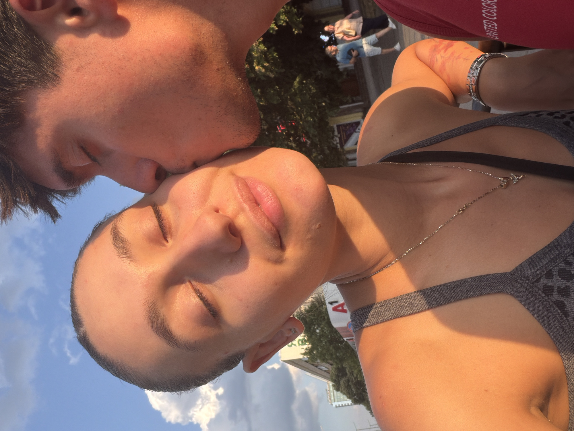 This is a close-up, selfie-style photograph taken outdoors during daylight, likely in an urban environment. A young woman is in the foreground, her eyes closed and her head tilted as a young man kisses her cheek. Both individuals are bathed in warm, golden sunlight, suggesting it's either early morning or late afternoon. The woman is wearing a grey sports bra and a silver necklace with a pendant. The man is wearing a red t-shirt.
In the background, out of focus, are buildings, trees, and a few other people walking. A building with a prominent "A1" logo is visible, which is a mobile network operator in Bulgaria, and is a common sight in Sofia. The sky above is blue with some white clouds. The overall impression is one of affection and a moment captured outdoors.
