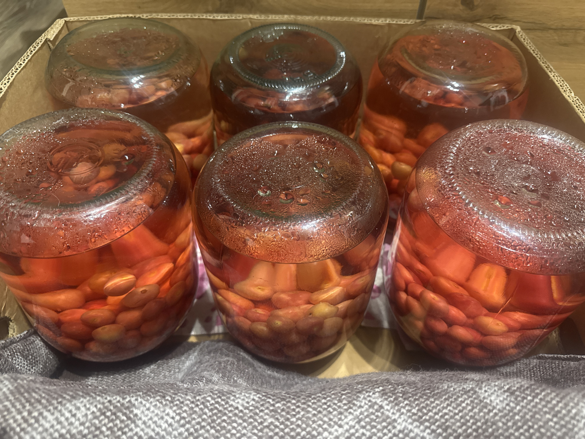 A close-up overhead shot shows six glass jars filled with red-colored liquid and what appear to be small, red fruits or berries. The jars are arranged in two rows of three, within a cardboard box. A grey, textured cloth lies at the bottom of the box, partially obscuring the lower portions of the jars. Condensation is visible on the inside of the glass jars, suggesting they may be cool or have recently been heated. The overall impression is that of preserved food items. The image's context is an undisclosed location within the city of Dobrovske settlement, Ukraine.