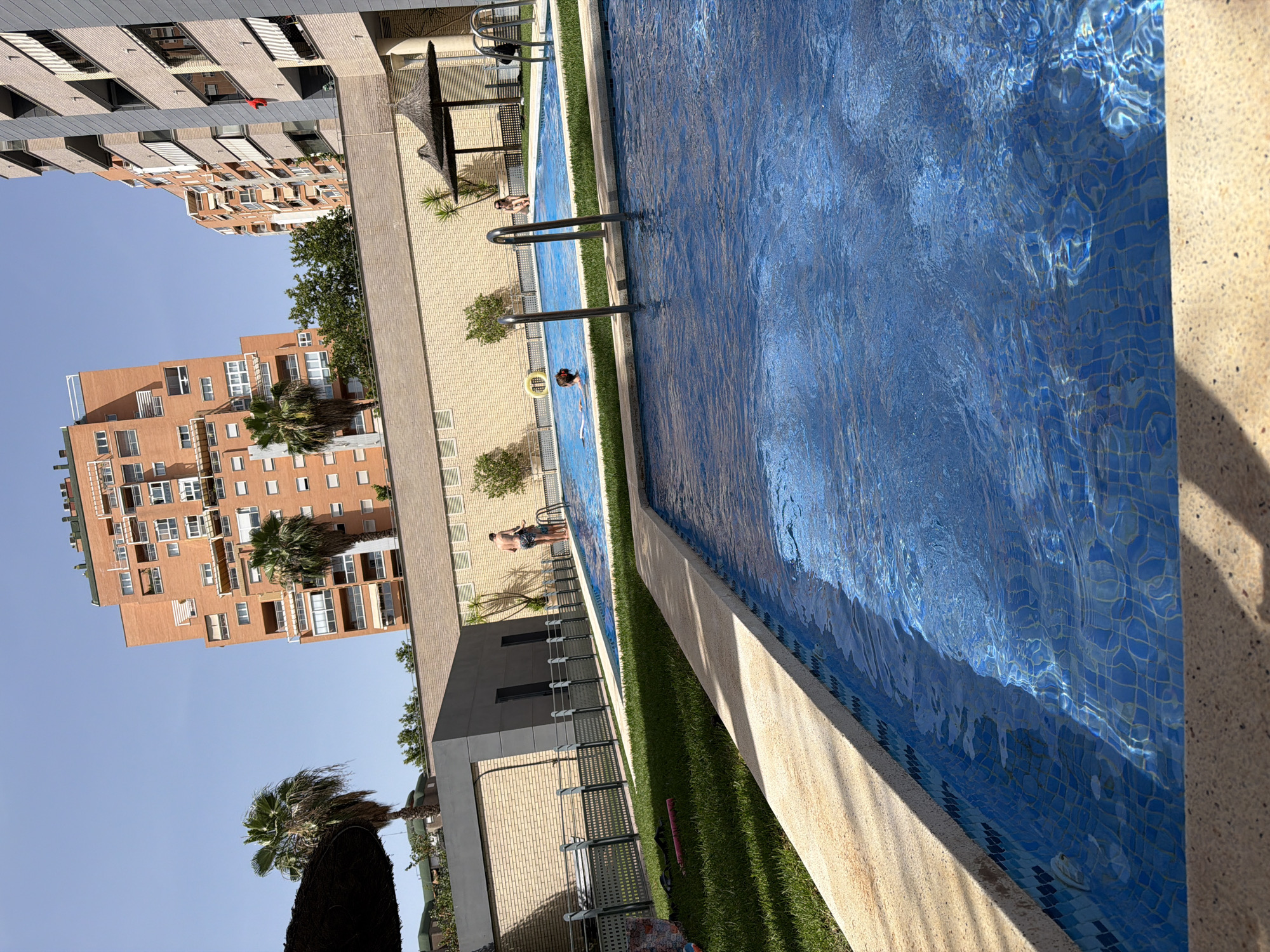 An outdoor scene depicts two swimming pools and surrounding residential buildings under a clear blue sky. In the foreground, a portion of a smaller swimming pool with blue tiled walls and a beige concrete coping is visible. The water is a vibrant blue, reflecting sunlight. Beyond this, a larger swimming pool with a yellow ring floating near the edge is in focus. Several people are present in and around the larger pool. Two individuals, one appearing to be an adult male and another a younger person, stand near the steps of the pool. A woman is seated on the edge of the larger pool under a thatched umbrella. In the background, several multi-story residential buildings with beige brick and white balconies dominate the skyline. Palm trees are scattered throughout the scene, and a section of manicured green lawn separates the pools from a low brick wall. The overall impression is one of a sunny, recreational environment in a developed urban area.