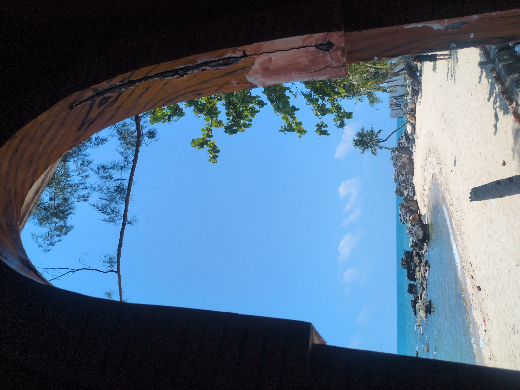 The image shows a beach scene framed by a damaged structure, possibly a building or part of a shelter, taken from a low angle. The frame consists of weathered wood, with some portions appearing to be painted. Through the opening, the beach is visible with white sand, a line of rocks along the shoreline, and the turquoise ocean. Vegetation, including palm trees and other foliage, can be seen. The sky is blue with scattered clouds. It is possibly located in the city of Phú Quốc, Vietnam.