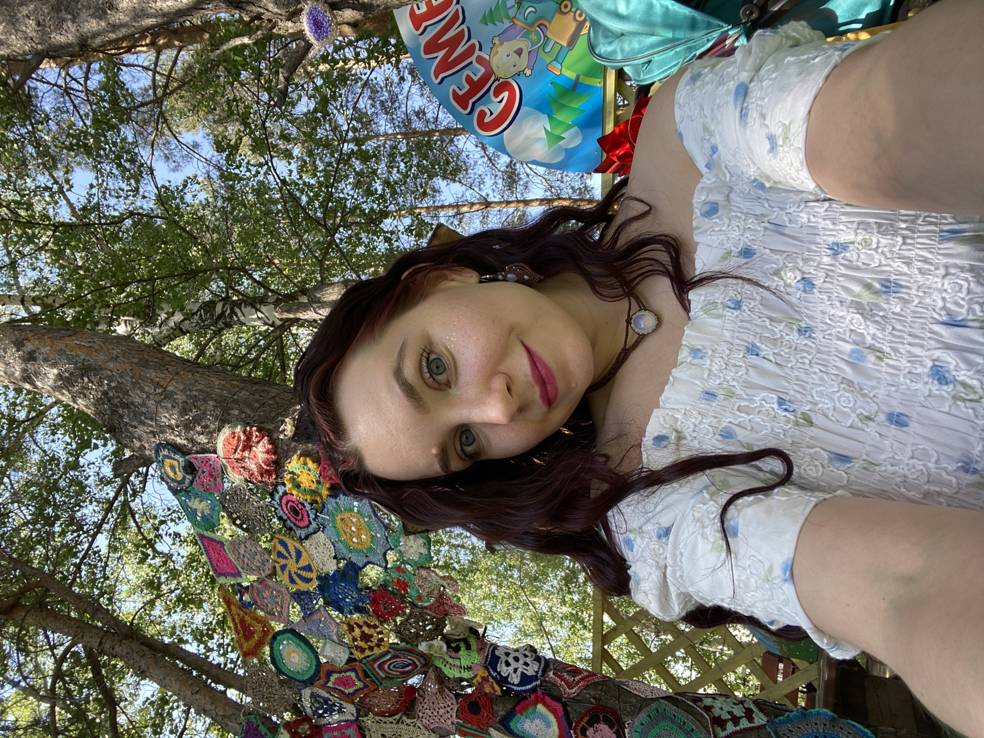 The image shows a woman with reddish-brown hair and green eyes looking into the camera. She is wearing a white dress with blue floral accents. The background features trees and greenery, suggesting an outdoor setting. A colorful, crocheted blanket hangs from the tree. A sign with "GEMA" is visible behind her. It is unknown if the setting is in Chaikovsky, Russia.