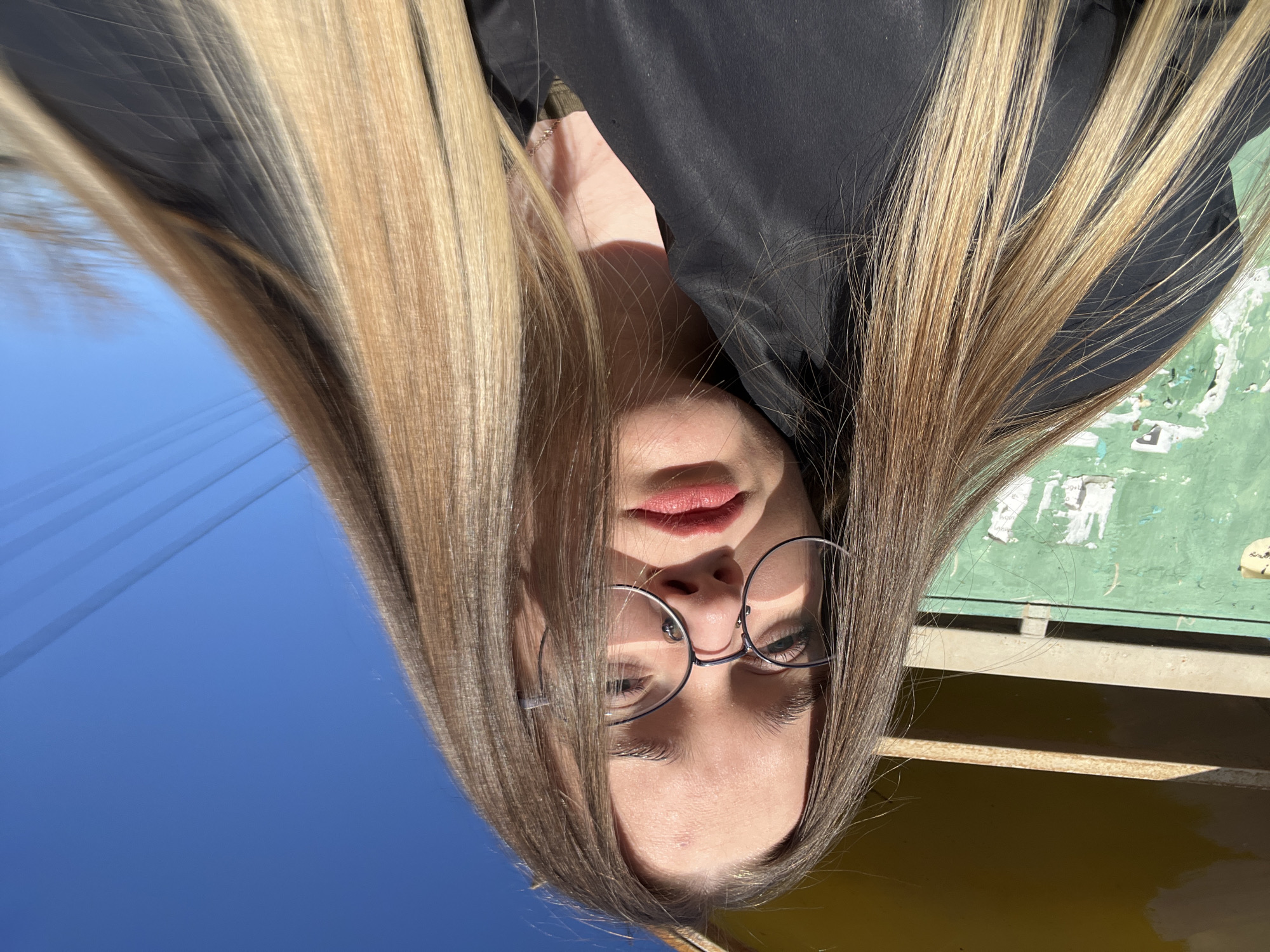 A close-up, low-angle self-portrait features a young female subject with light-colored, long, straight hair, styled with an ombre effect transitioning from brown at the roots to blonde at the ends. She wears round, silver-rimmed eyeglasses and a black collared jacket over what appears to be a dark green shirt. Her lips are covered with a red lipstick, and a faint mark is visible on her forehead. She maintains direct eye contact with the camera. The background on the left consists of a weathered, pale green wall with peeling paint and remnants of white paper affixed to it, topped by a section of a yellow structure. To the right and top, a clear, bright blue sky is visible, traversed by several thin, dark horizontal lines, consistent with overhead wires. The scene is illuminated by direct sunlight, casting shadows on the subject's face. The image depicts an outdoor urban setting in the city of Большевик, Russia.