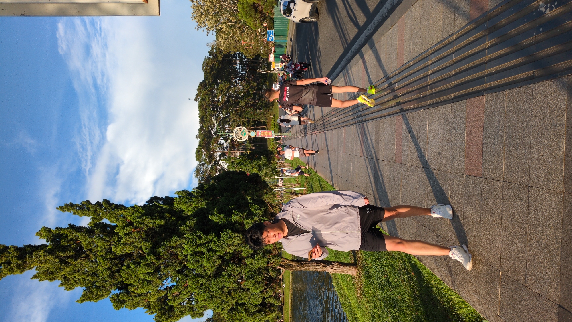 In Da Lat, Vietnam, a young Asian man in a light grey jacket and black shorts walks along a paved sidewalk beside a body of water. He is looking down at a mobile phone in his hand. Several other individuals, dressed in athletic wear, are visible further down the sidewalk, some appearing to be running or jogging. Behind the sidewalk is a grassy area and a tall, dense green tree, characteristic of Da Lat's cooler climate. Beyond the trees, a road can be seen with vehicles, including a white car and motorcycles. The sky above is blue with scattered white clouds, suggesting it is daytime. The overall scene depicts a casual outdoor activity in a park-like setting within the city.