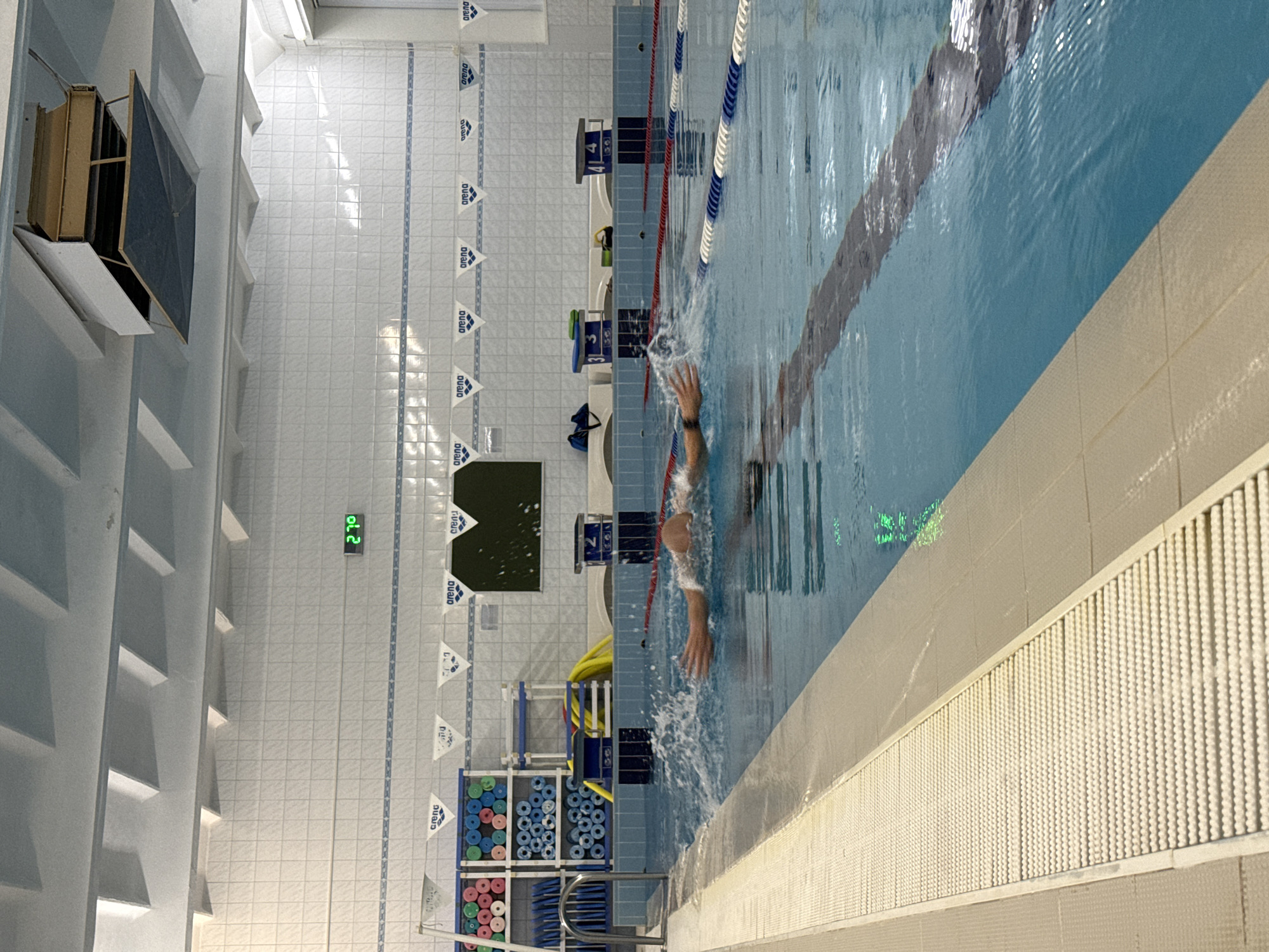 This is an indoor swimming pool. A person is swimming in the pool, which is divided into lanes by lane markers. The lane markers are white and blue. The pool edge is white with a textured surface. There are starting blocks with lane numbers (4, 3, 2) along one side. The pool is marked with flags bearing the brand "Arena." Wall shelves with blue foam rollers and pool equipment are visible near the pool’s edge. A digital display on the wall shows "27.0." The tiles are white with blue decorative stripes.
Based on visual cues such as the interior design, pool markings, and overall appearance, this swimming pool could potentially be located in the city of Chaikovsky, Russia.