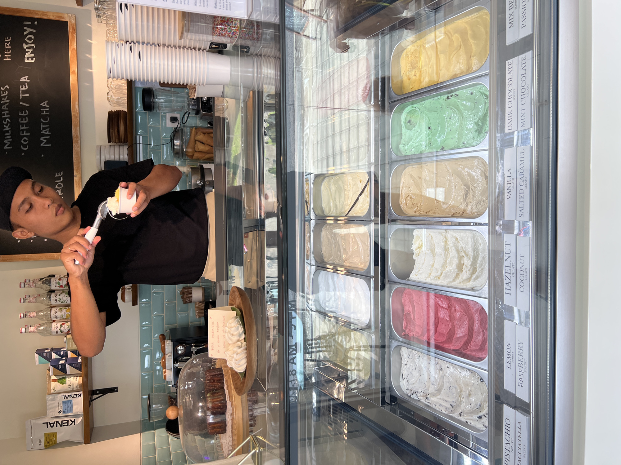 An indoor shot captures a gelato shop counter in Abang, Indonesia. A young man wearing a black t-shirt and black cap is scooping gelato into a white cup with a metal scoop. The gelato counter displays various flavors in stainless steel trays. Behind the man, shelves are stocked with bottles, boxes, and cups. The environment is clean and well-lit, with a tiled backsplash and a wooden menu board. The overall scene suggests a typical bustling gelato shop.