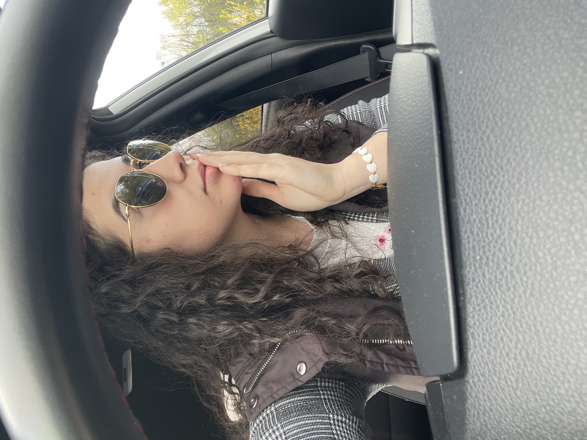 The image captures a close-up portrait of a young woman from inside a car, rotated 90 degrees counter-clockwise. She has long, dark, curly hair and is wearing gold-framed aviator sunglasses that reflect a bright, sunlit sky and green trees. Her left hand is raised to her face, with light-colored fingernails resting near her mouth and chin. On her wrist, she wears a bracelet composed of white, heart-shaped beads.
Her attire includes a black-and-white plaid top or jacket, layered under what appears to be a dark brown or black quilted vest with metallic snap buttons. The car's interior is visible, including a section of the steering wheel on the left with red stitching, a black textured dashboard to her right, and a black seatbelt mechanism in the background. Bright daylight floods the scene from above and behind her, indicating it is daytime with sunny weather. The background outside the car shows glimpses of green foliage, suggesting a natural outdoor setting. Her pose is contemplative or stylish. There is no visible text in the image. The location context provided is Sofia, Bulgaria.