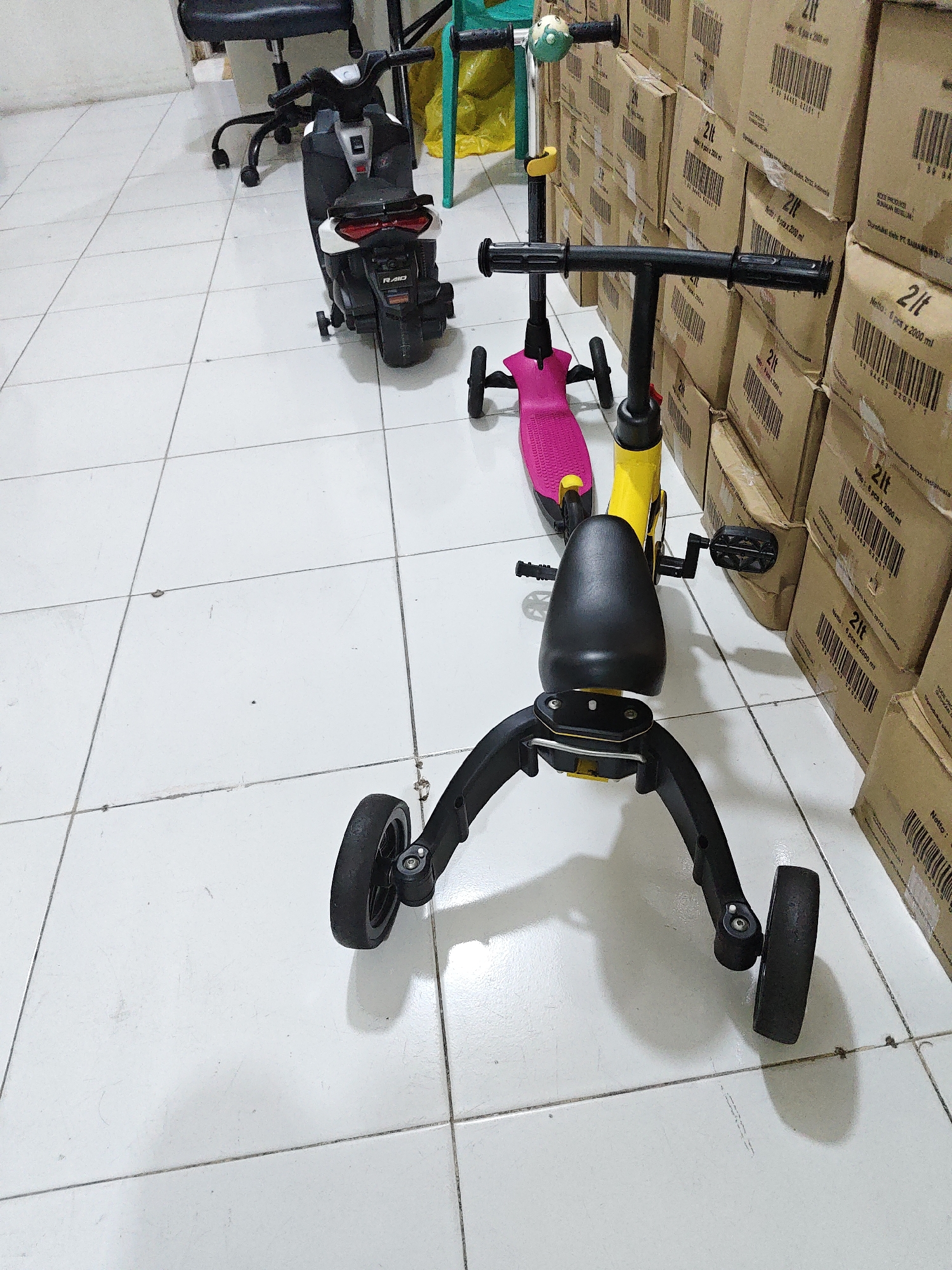 The image depicts a storage area in North Jakarta, Indonesia, likely a warehouse or a large retail backroom, given the stacks of cardboard boxes in the background. The floor is tiled with white square tiles, and the lighting suggests an indoor environment.
In the foreground, a multi-functional child's ride-on toy is the primary subject. It appears to be a 3-in-1 model, capable of functioning as a scooter, a tricycle, or a balance bike. The ride-on toy is primarily black and yellow, with a pink deck. It has three wheels: two smaller ones at the front and a single larger one at the back. The handlebars are black and feature black grips. The seat is black and ergonomically shaped. The pedals are visible and are black.
To the left and slightly behind the ride-on toy is a black electric toy motorcycle. This toy also has a seat, handlebars, and wheels. It is predominantly black with white accents and red taillights. A portion of a black office chair with wheels is visible to the far left.
Behind the ride-on toys, a stack of cardboard boxes is prominently displayed. The boxes are light brown and appear to contain products, possibly liquids, given the "2lt" labeling and bar codes visible on some of them. Some of the boxes also have text that seems to be in Indonesian. A green plastic chair with a yellow cushion is partially visible between the toy motorcycle and the stacks of boxes. A yellow plastic bag or sheeting is also present in the background, draped over a structure. The overall impression is one of a commercial space used for storing and possibly displaying children's toys and other merchandise.