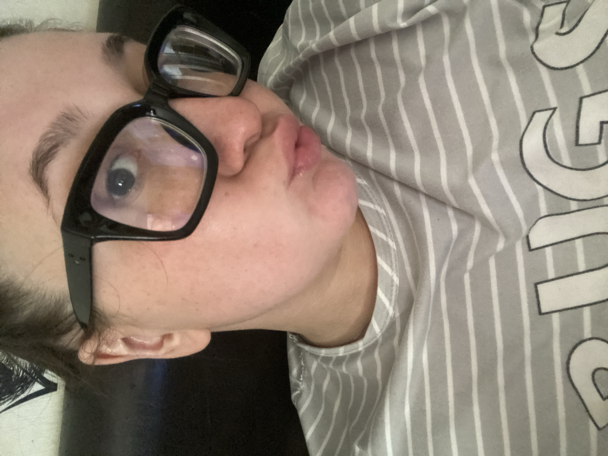 The image shows a person wearing black-framed glasses and a grey and white striped shirt. The person is positioned at an angle, with the head tilted to the side. The shirt has white lettering, some of which is visible. No details suggest the location is Chaikovsky, Russia.