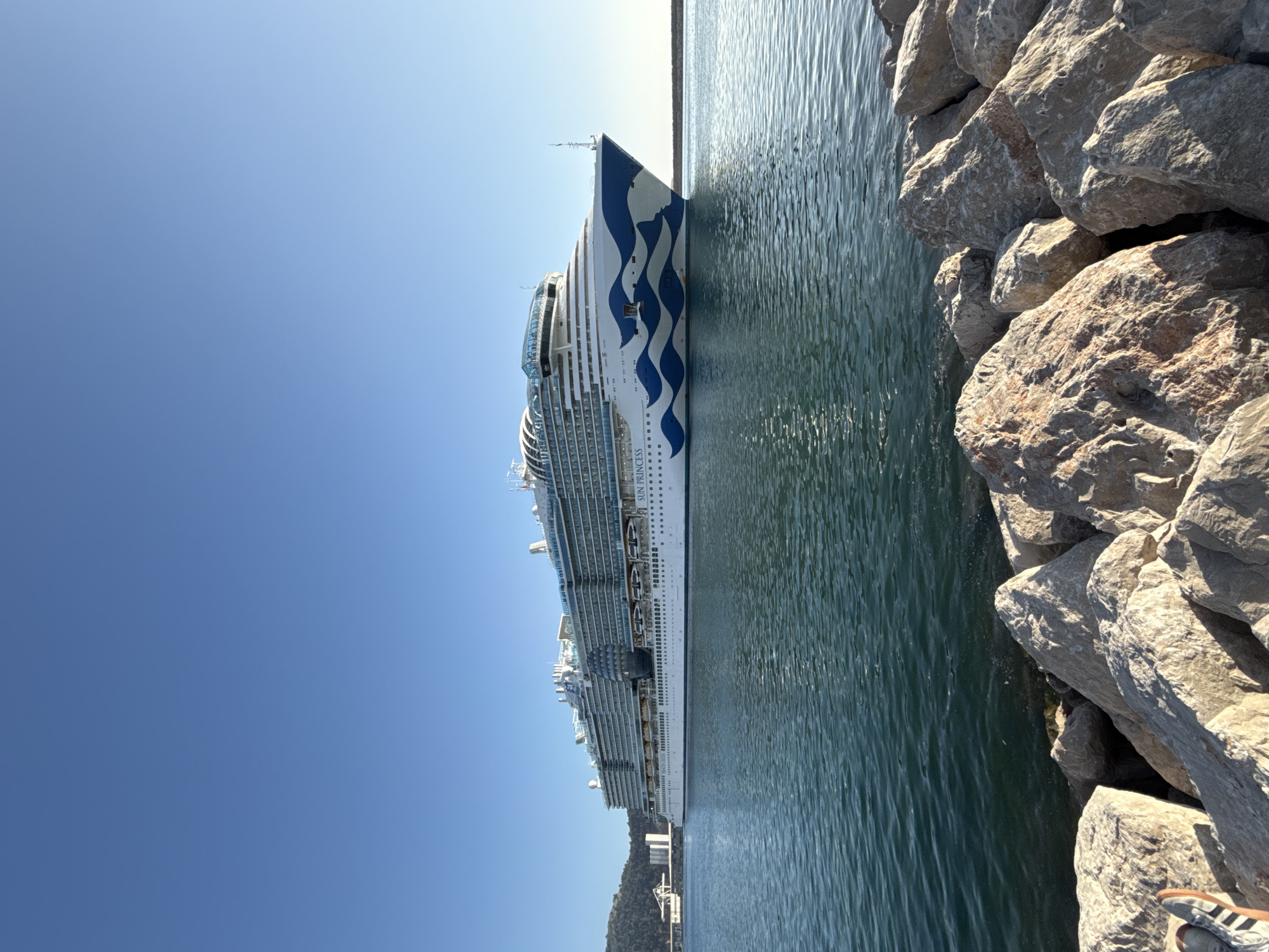 A large cruise ship is docked next to a rocky breakwater in Bar, Montenegro. The ship is painted white with blue wave designs. The sea is calm, and the sky is clear blue. The breakwater is made of large, gray rocks. A foot wearing a sandal is visible in the lower right corner of the frame. In the background, a building is seen at the foot of a mountain.