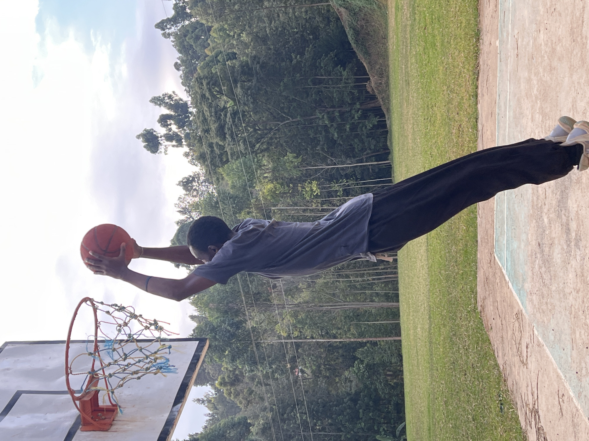 A young Black male wearing a grey t-shirt and black pants is captured mid-air, dunking a basketball into a hoop. He is on an outdoor basketball court with a grassy field and a densely wooded hillside in the background. The sky is partly cloudy. The basketball hoop is attached to a white backboard. The court appears to be made of concrete with painted lines. This scene is located in Kamakwa/Mukaro Ward, Kenya.