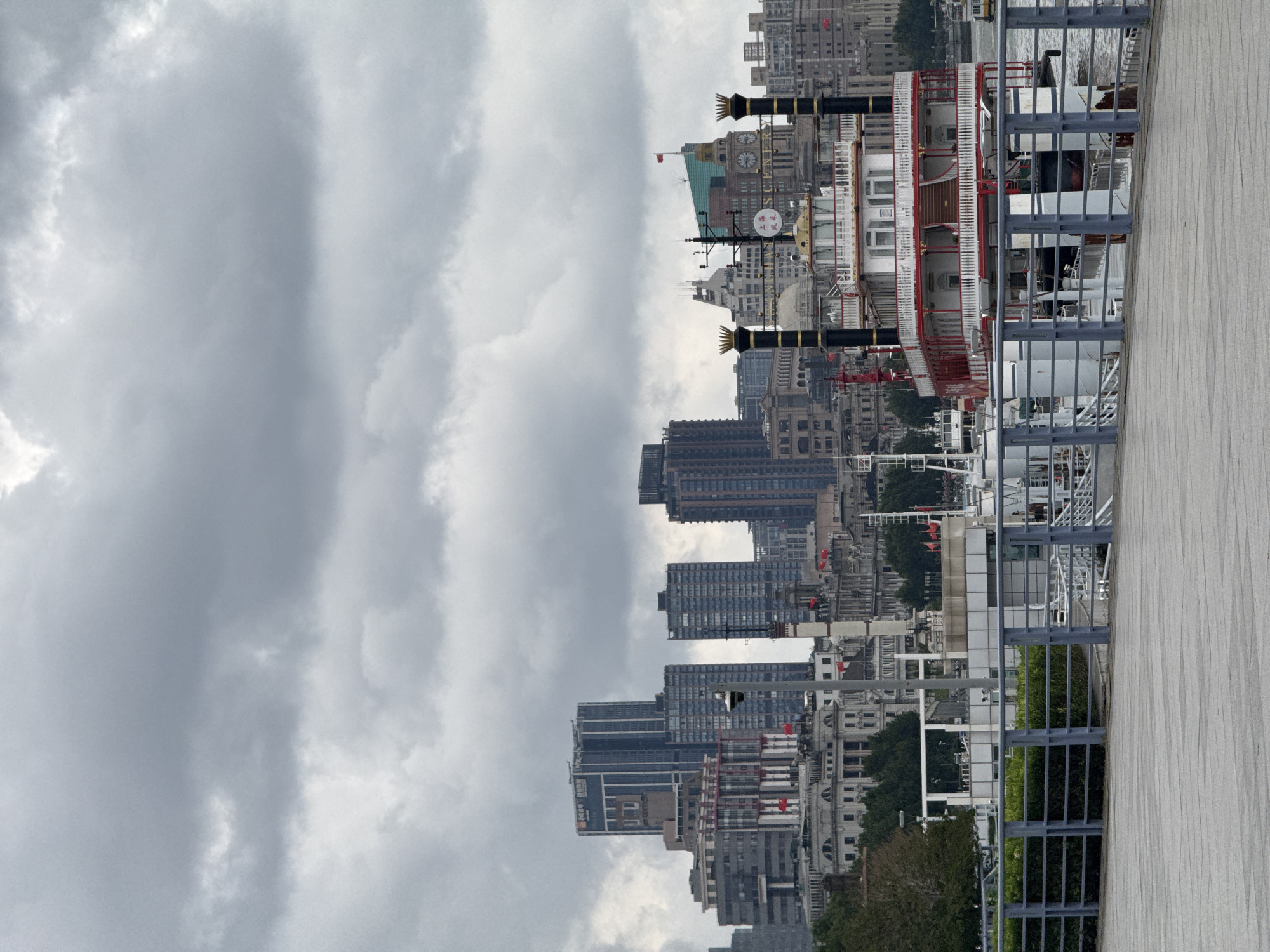 The image depicts a cloudy day in Shanghai, China, along the Bund waterfront. In the foreground, a wooden boardwalk runs horizontally across the bottom of the frame, separated from the water by a blue metal railing. Beyond the railing, a large, multi-decked riverboat with a white and red exterior and a paddlewheel structure is docked. Several other boats are visible in the water behind it. The middle ground features a collection of varied architectural styles, including historic European-style buildings with classical facades and taller, modern skyscrapers piercing the overcast sky. Red Chinese flags are visible on some of the older buildings. The background is dominated by the dense urban skyline of Shanghai, characterized by numerous tall buildings of different shapes and colors, all under a heavy blanket of gray clouds. The overall atmosphere is muted and slightly somber due to the weather. There are no visible people in the frame.