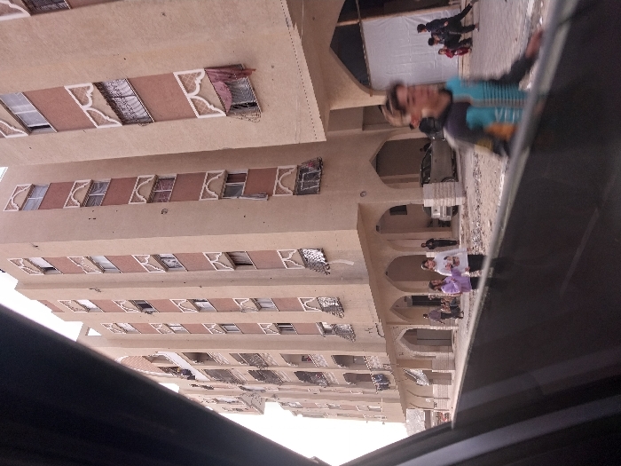 A multi-story building with ornate windows and arched walkways is captured from a low angle, presumably from within a vehicle. People are visible walking along the street below the building's archways. A person wearing a blue and black shirt is also seen. The vehicle's interior is partially visible in the foreground.