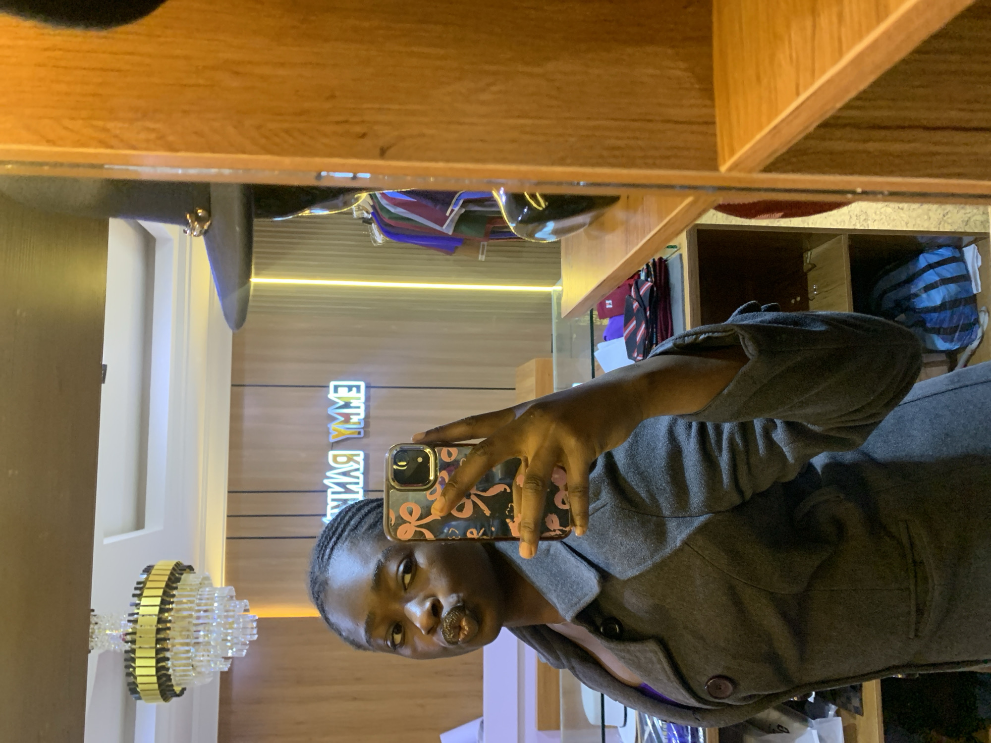 A young Black woman with braided hair poses for a selfie in what appears to be a boutique or store. She is wearing a dark grey coat over a purple garment and is holding a smartphone in her right hand. The phone case has a pink bow pattern. The background shows a store interior with wooden shelving, a chandelier, and a neon sign that reads "EMMY BANK" in reverse. Clothing racks with various garments are also visible, along with a black hat on display. The overall impression is a casual, everyday moment captured in a retail setting.