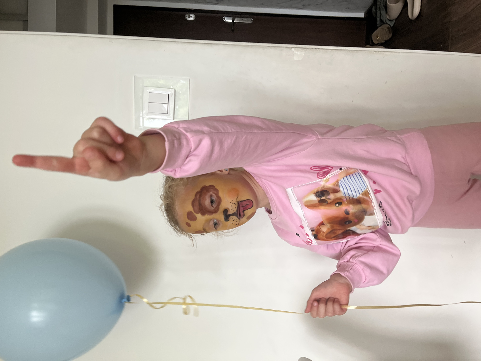 A young child with dog face paint is holding a light blue balloon with a gold ribbon. The child is wearing a pink long-sleeved shirt with a dog print on the front. The child is extending their index finger. In the background is a white wall with a light switch and a dark wooden door. The location is Unknown, Unknown.