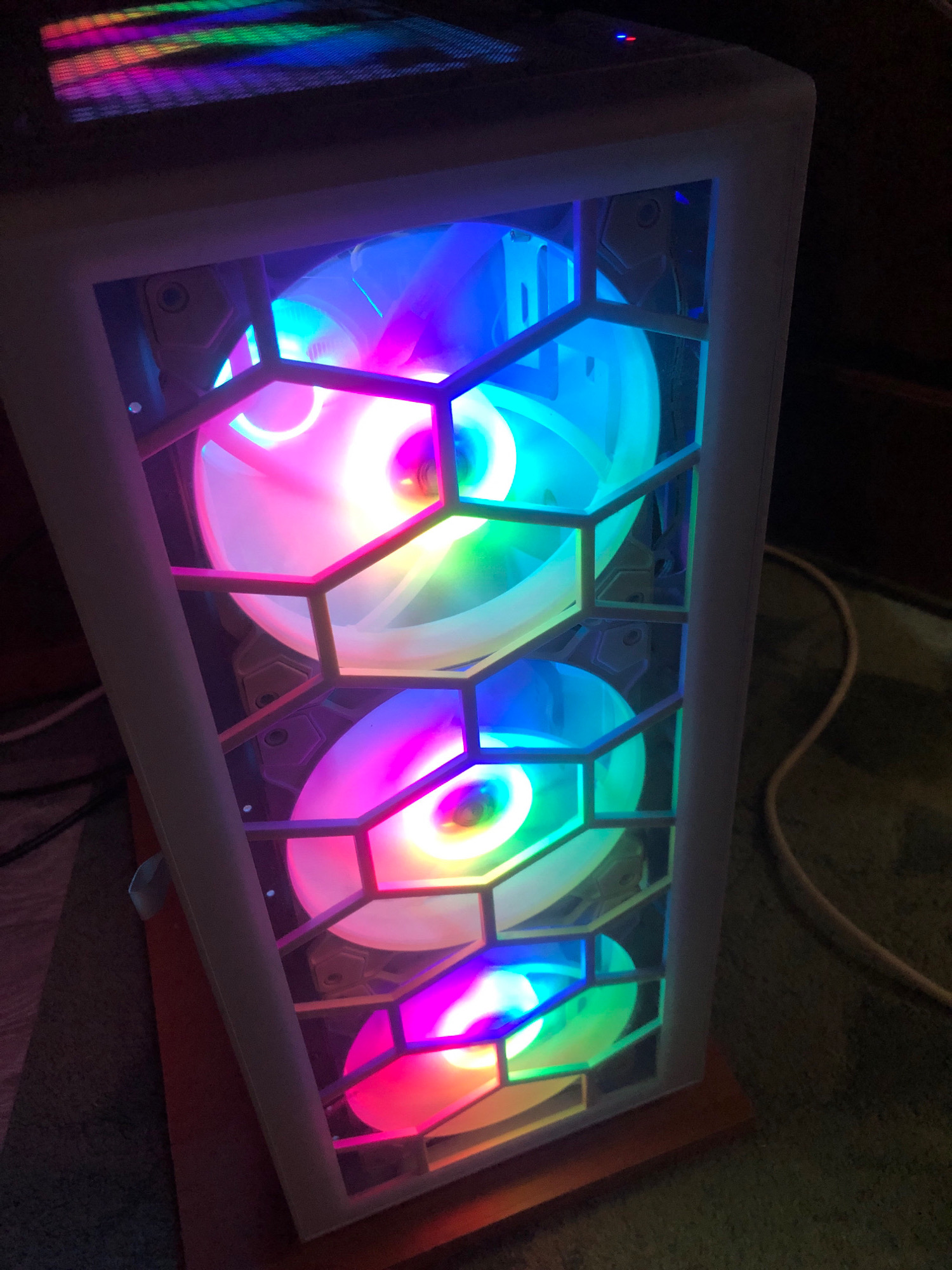 The image shows a white computer tower with multiple RGB fans that are illuminated with a rainbow of colors. The fans are visible through a hexagonal mesh grille on the front of the tower. The computer is placed on a wooden surface, possibly a desk or shelf, and there are cables visible in the background. The lighting in the image is dim, with the primary illumination coming from the computer's internal lights. There are no people or other objects of note in the immediate vicinity of the computer. The background appears to be a domestic setting, likely a room. The image does not contain any information that would suggest a location in Borisovka, Russia, or any other specific geographical location.