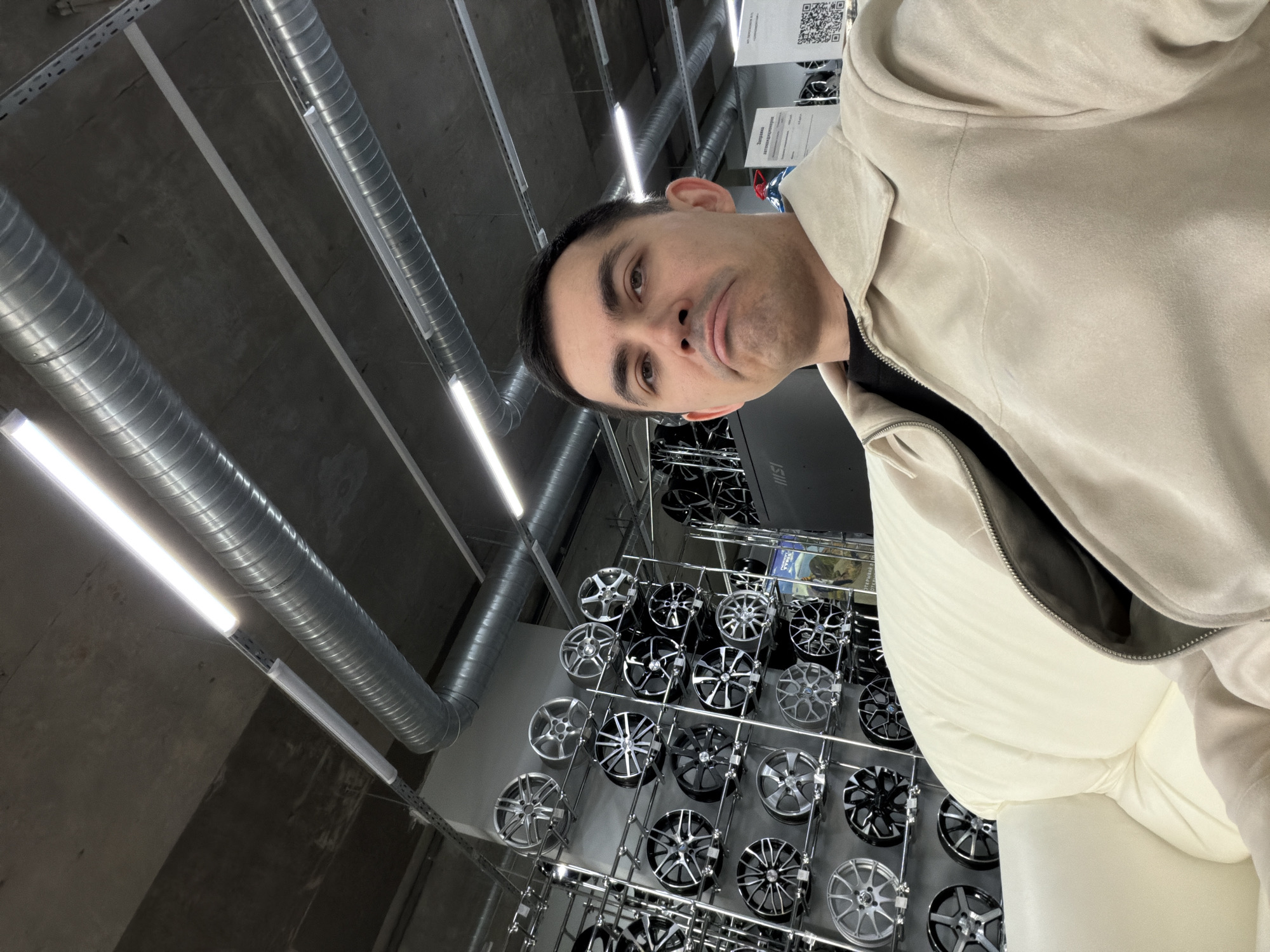 A man stands in a store, likely in Izhevsk, Russia, as suggested by Cyrillic text on displays. He wears a light-colored hooded jacket. Behind him, a wall displays numerous car wheels organized on metal shelving. The environment suggests an industrial or retail setting. The ceiling has exposed ductwork and fluorescent lighting.