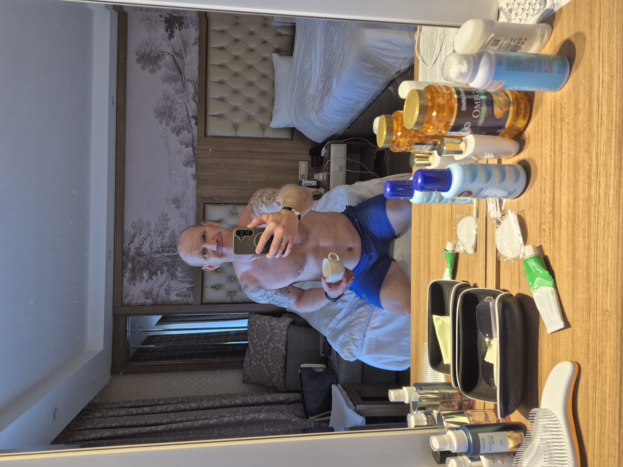 A shirtless, balding man with a tattoo on his left arm takes a selfie in a hotel room mirror in Alanya, Turkey. He is wearing blue shorts and holding a small white cup. Reflected in the mirror is a bed with a headboard featuring a landscape print, and a bedside table. In the foreground, on a wooden surface, are several bottles of toiletries, a comb, and a sunglasses case. The room's decor includes patterned curtains and what appear to be additional seating.