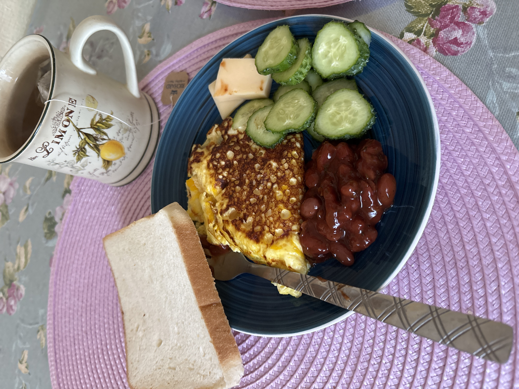 This is a close-up, top-down shot of a breakfast meal served on a dark blue plate, placed on a pink woven placemat. The meal consists of an omelet, baked beans, sliced cucumbers, and a few cubes of cheese. A slice of white bread is positioned on the top left of the plate. A fork with a patterned handle rests on the left side of the plate, partially submerged in the omelet. To the right of the plate, a mug filled with tea sits on the placemat, with a tea bag tag visible. The background features a floral-patterned tablecloth. The scene is a tabletop setting, likely indoors, and there is no indication of the city of Naberezhnye Chelny, Russia. The image focuses solely on the food and table setting.
