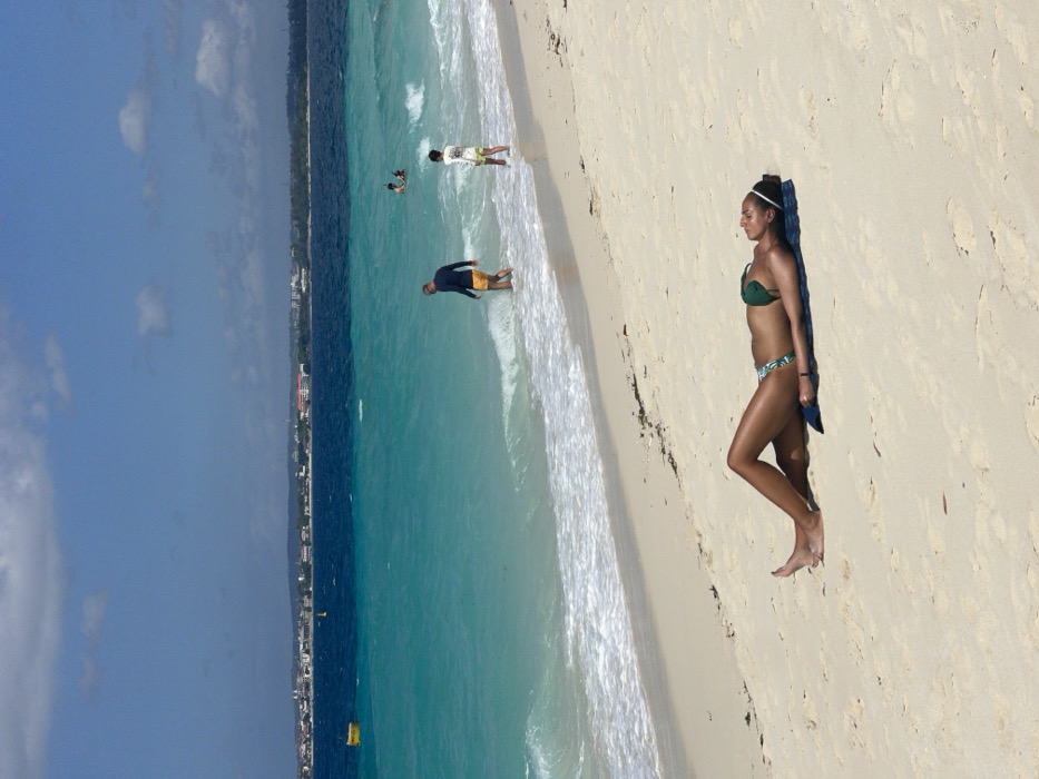 The image displays a sunny beach scene located in the city of Zanzibar, Tanzania, facing a body of water identified as the Indian Ocean. In the immediate foreground, a female individual with tanned skin is lying on her back on a dark blue mat placed directly on the light beige sand. She is wearing a dark green bikini top and patterned bikini bottoms. Her arms are positioned alongside her body, and her legs are extended. A white headband is visible in her hair.
Moving towards the water, patches of dark seaweed or debris are present on the sand near the waterline. In the midground, two male individuals are situated at the water's edge. One man, wearing a dark long-sleeved rash guard and yellow swim shorts, is wading into the turquoise water, facing away from the camera. The second man, dressed in a light-colored t-shirt with a graphic and dark shorts, stands in the shallow surf nearby. Further out in the turquoise water, two additional individuals are partially submerged.
The water transitions from a clear turquoise near the shore, where small white waves are breaking, to a deeper blue further from the coastline. A yellow cylindrical buoy is visible floating in the deeper blue water to the left side of the frame. Across the expanse of water, a distinct distant cityscape composed of various light-colored buildings lines the horizon, confirming an urban coastal setting. The sky above is clear blue with scattered white cumulus clouds.