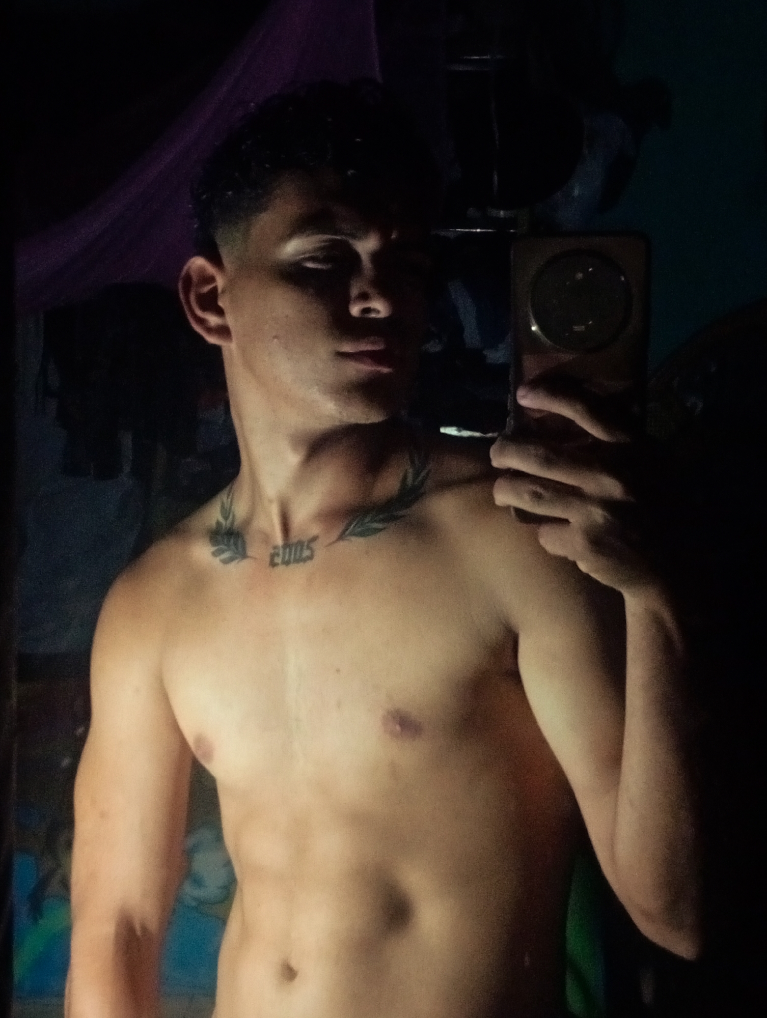 A shirtless young man with short dark hair and a tattoo on his upper chest/collarbone area that says "2005" is taking a selfie with a cell phone. The lighting is dim. There is a partially visible room in the background. There is no information in the image to indicate that this was taken in the city of Altagracia, Venezuela.