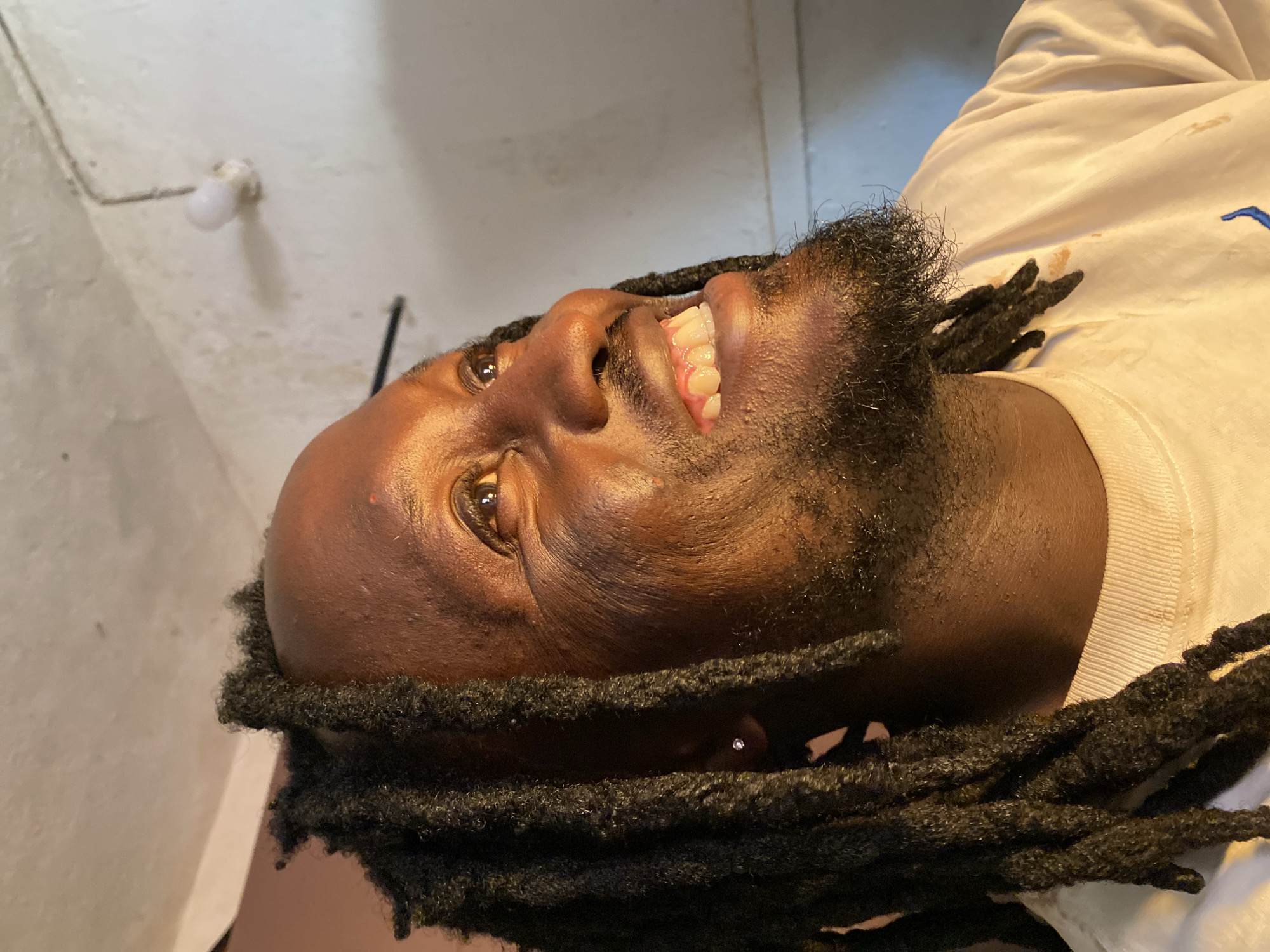 An upward-facing close-up shot captures a dark-skinned man smiling widely, revealing his upper teeth. He has a full beard and dark dreadlocks framing his face and extending to his shoulders. A small, shiny earring is visible in his left ear. He appears joyful or amused, with his eyes looking towards the upper left of the frame.
He is wearing a white, long-sleeved top, possibly a t-shirt or sweatshirt, which has faint stains and a partial blue embroidered design (resembling a Nike swoosh) on the right side.
The background consists of a plain, light-colored (off-white or greyish) wall or ceiling, which shows some texture and appears somewhat aged or worn. An old-fashioned, small white light bulb in a simple fixture is attached to the wall/ceiling in the upper left, with an electrical wire running past it. The lighting is somewhat dim and uneven, suggesting an indoor setting. There is no discernible weather, but the artificial light source indicates it could be any time of day in a space that requires illumination. The overall impression is informal and candid. No text is visible in the image. The setting is in Vom, Nigeria.