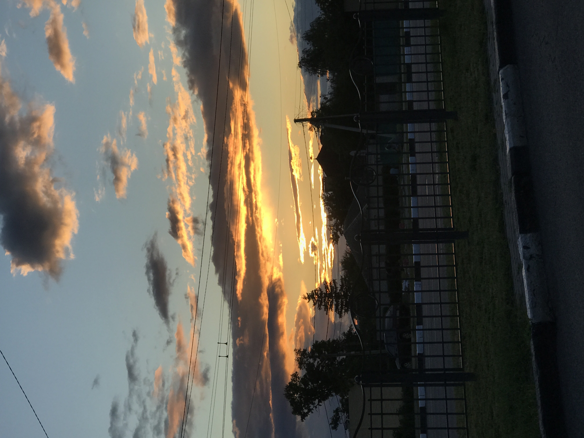 The image depicts a sunset sky in an urban setting. The sky is filled with clouds illuminated with orange and yellow hues of the setting sun, while some clouds are darker. Electrical wires stretch across the sky. The view includes a fence and some trees. A street curb is visible at the edge of the frame. The location is Unknown.