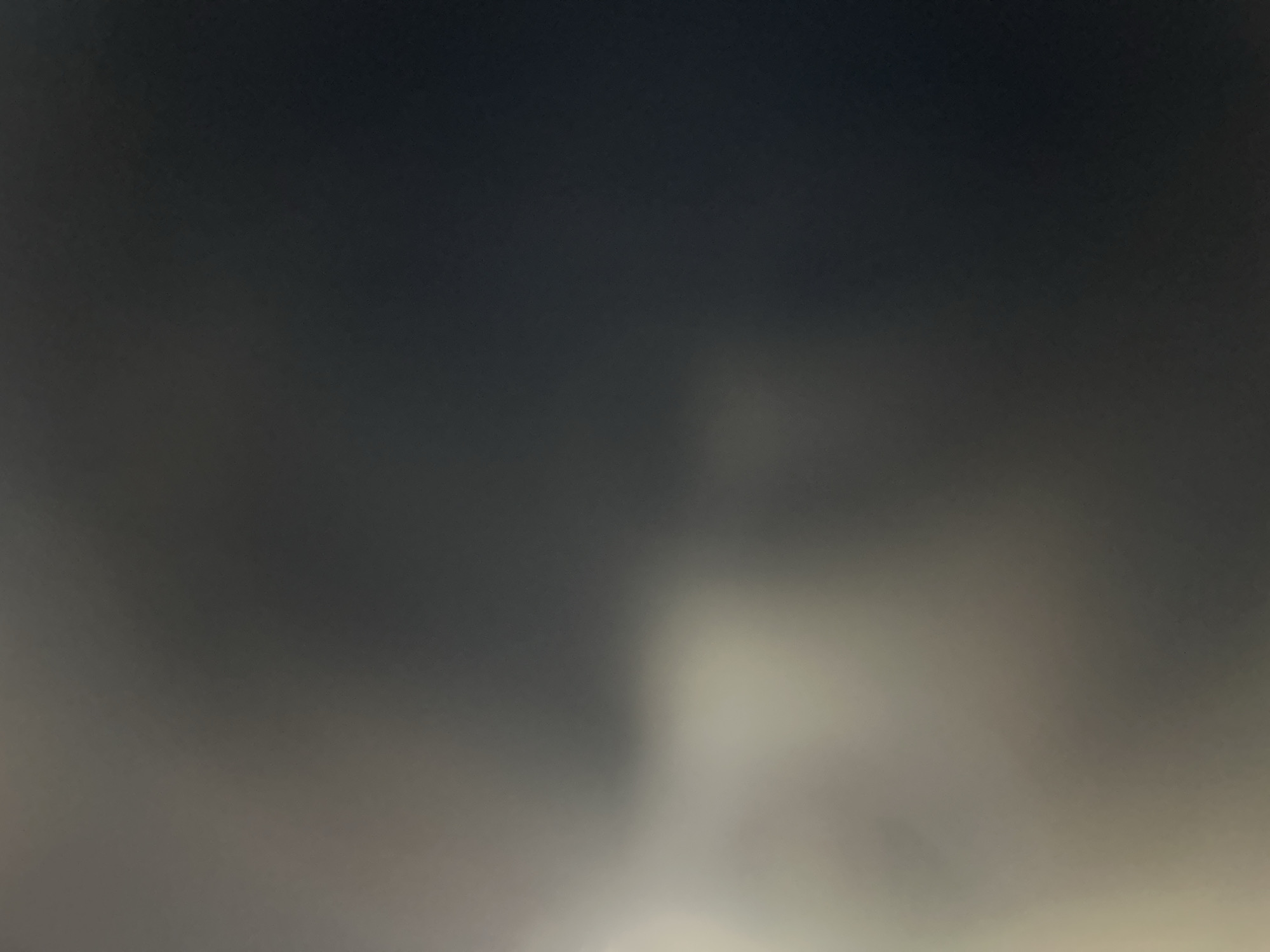 The media file is a dark, abstract image with soft, diffused lighting. The dominant colors are shades of gray, ranging from very dark in the upper portion to lighter, almost white tones in the lower half. There are no discernible subjects such as people, objects, or a clear scene. The lighting creates a luminous effect in the lower part of the image, suggesting a source of light outside the frame or a very hazy atmosphere. The texture is smooth and uniform, with no sharp edges or distinct features. The overall impression is one of a dark, possibly overcast sky or a dimly lit, foggy environment. There is no visible text. Given the location context of Belgrade, Serbia, this could potentially depict a foggy night sky over the city, or a heavily overcast day with diffused light. The abstract nature of the image makes it difficult to determine a specific activity or event, or to infer emotions beyond a general sense of subdued or atmospheric lighting.