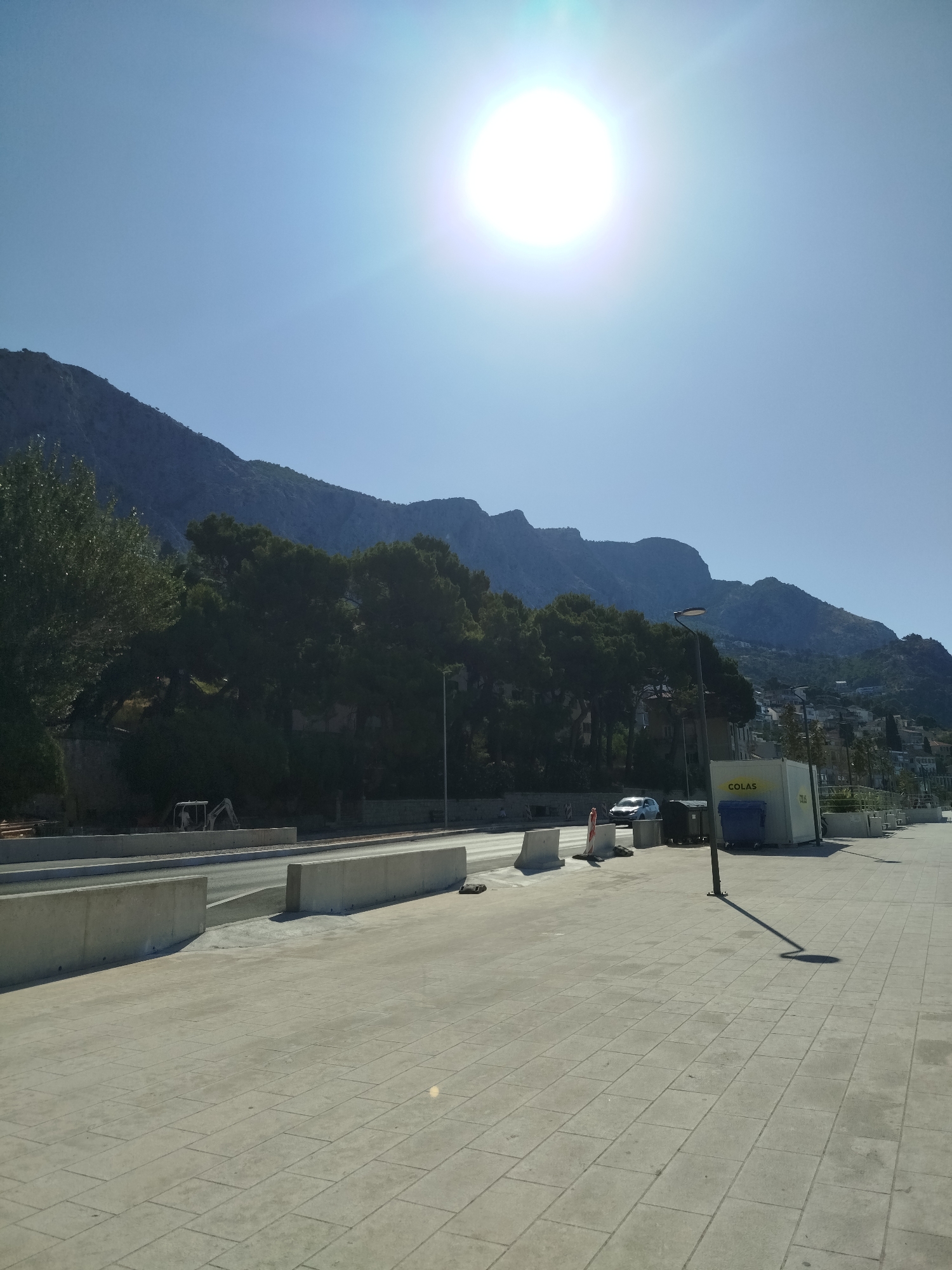 The image shows a sunny day in Omiš, Croatia, with a clear blue sky and the sun prominently displayed in the upper center, casting a lens flare. In the foreground, a paved walkway made of light-colored rectangular tiles stretches horizontally across the frame. To the left, there is a construction site with concrete barriers and an excavator partially visible. Beyond the construction, a road is visible with a white car driving on it. A line of dark green trees, likely pines, borders the road and the construction area. Behind the trees, a large mountain range dominates the background, with some buildings and structures scattered on its slopes. A tall street lamp with a single light fixture casts a long shadow on the walkway. To the right of the street lamp, there is a white shipping container with the text "COLAS" and a dark blue dumpster. The overall scene suggests an urban or semi-urban environment adjacent to a natural mountainous landscape.