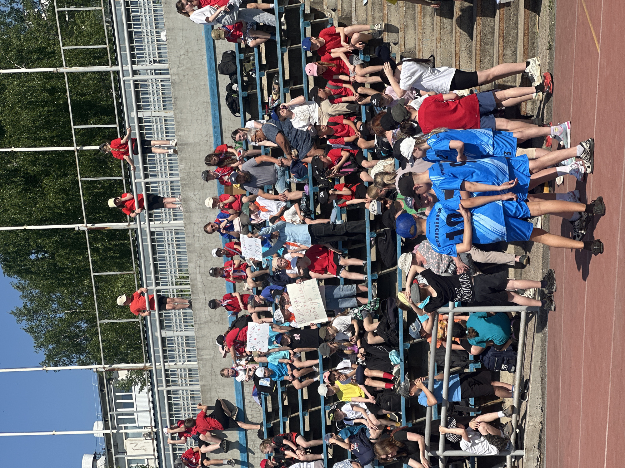 A crowd of people is seated in the bleachers of a stadium in Votkinsk, Russia. Most are wearing red or blue shirts, with some holding up signs written in Russian. The environment is sunny. A track runs along the front of the bleachers.