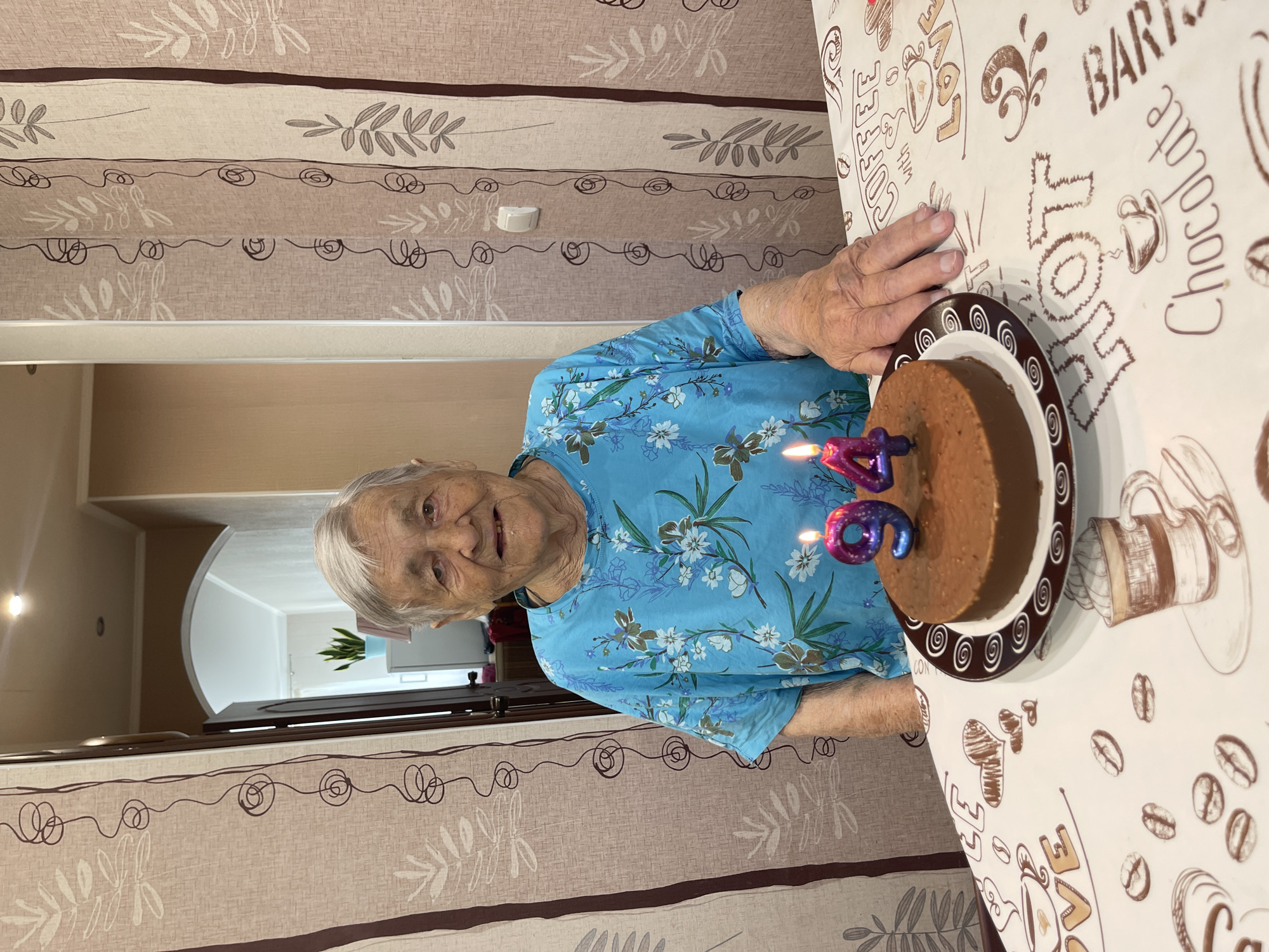 An elderly woman with gray hair, wearing a blue floral shirt, smiles at the camera. She is seated at a table with a chocolate cake in front of her, adorned with two candles shaped like the numbers '9' and '4'. The table is covered with a tablecloth featuring coffee-themed illustrations. The background shows a doorway leading to another room and patterned curtains. The scene appears to be an indoor celebration.