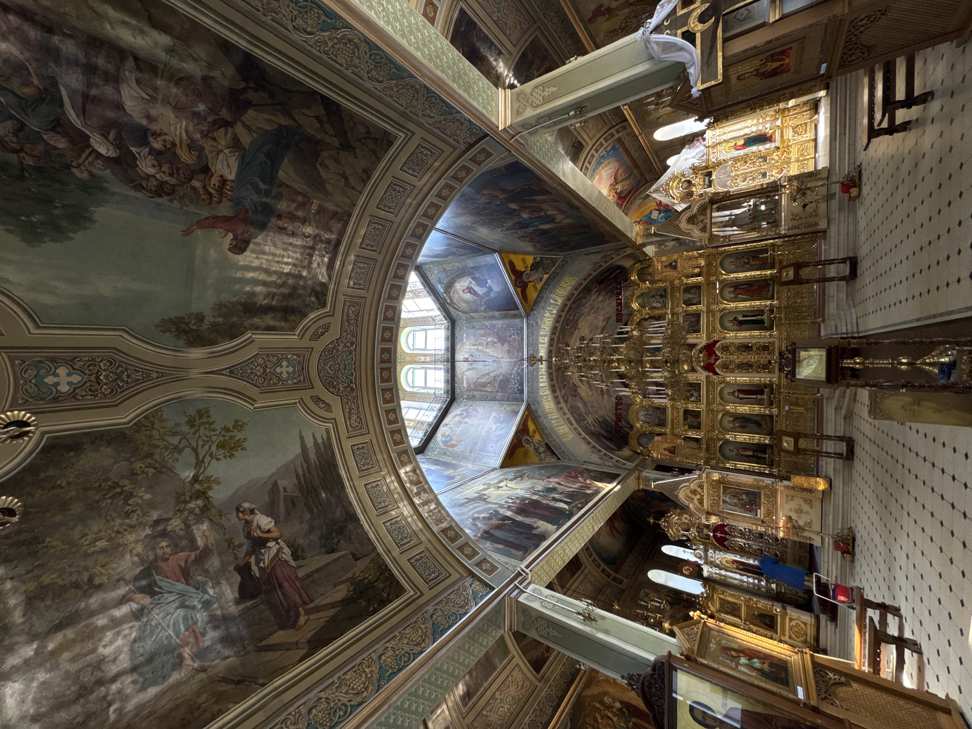 The image depicts the interior of an Orthodox church, showcasing a richly decorated space with ornate golden iconostasis and extensive ceiling frescoes. The perspective is wide-angle, capturing the grandeur of the architecture and the religious art. In the foreground, the tiled floor leads towards the altar area. The walls and ceiling are adorned with numerous painted scenes, likely biblical narratives, in a traditional style. Light streams in from windows, illuminating parts of the interior and creating a dramatic effect. Several large icons are displayed on wooden stands in the left foreground. The overall impression is one of opulence, spirituality, and historical significance. No people are clearly visible in the scene, though some figures are depicted within the paintings on the ceiling and walls.
