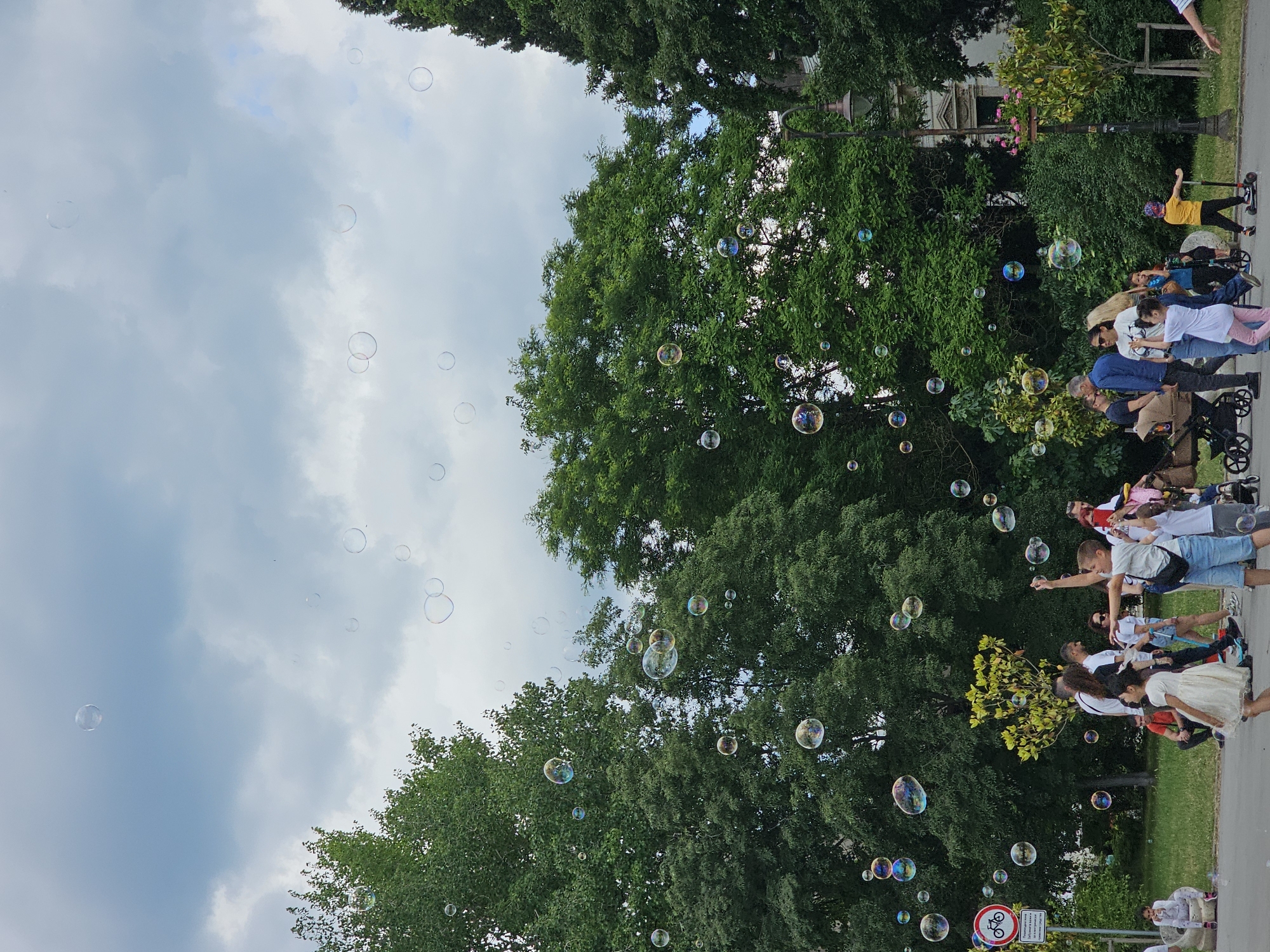 The image depicts a group of people in an outdoor setting, possibly a park, with numerous bubbles floating in the air. Green trees form a backdrop, interspersed with views of a cloudy sky.
On the right side of the image, a group of people stands or moves along a pathway. Individuals include children, adults, and those pushing strollers, some of whom seem to be interacting with the bubbles. There is a person on a scooter.
In the lower right corner of the frame is a street sign with a bicycle on it. The location seems to be within an urban area given the presence of the street sign. The city and country are unknown.