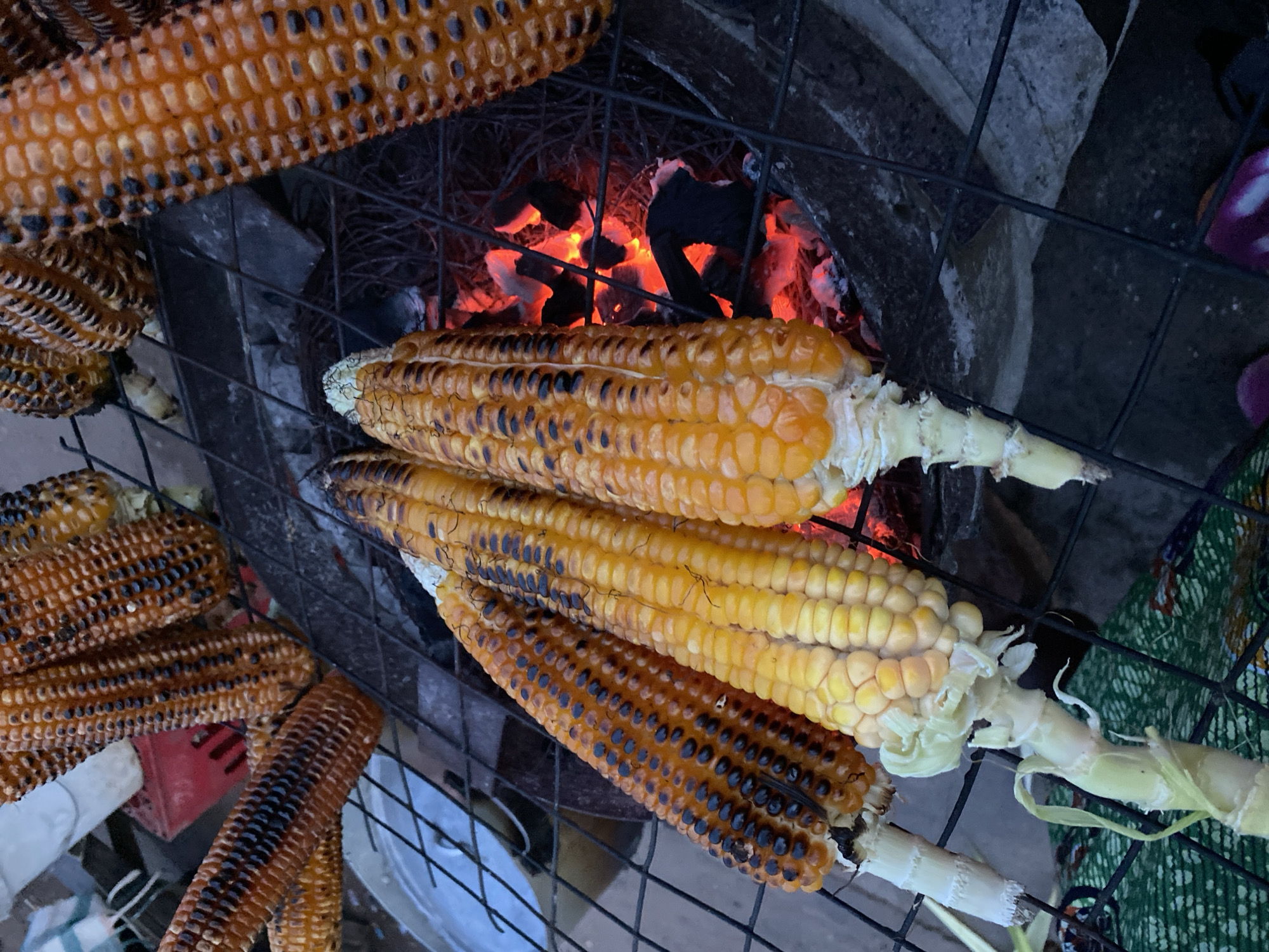 The image shows an outdoor cooking scene in Bukuru, Nigeria, featuring several ears of corn being grilled over hot coals. The corn cobs are yellow and some have been charred to varying degrees, with visible black spots and stripes on their kernels. They are arranged on a black wire grill, positioned above glowing red embers. The background is dark, suggesting it is either evening or the area is shaded. In the upper left, a red plastic crate is partially visible. The overall impression is of a rustic, informal outdoor cooking setup.