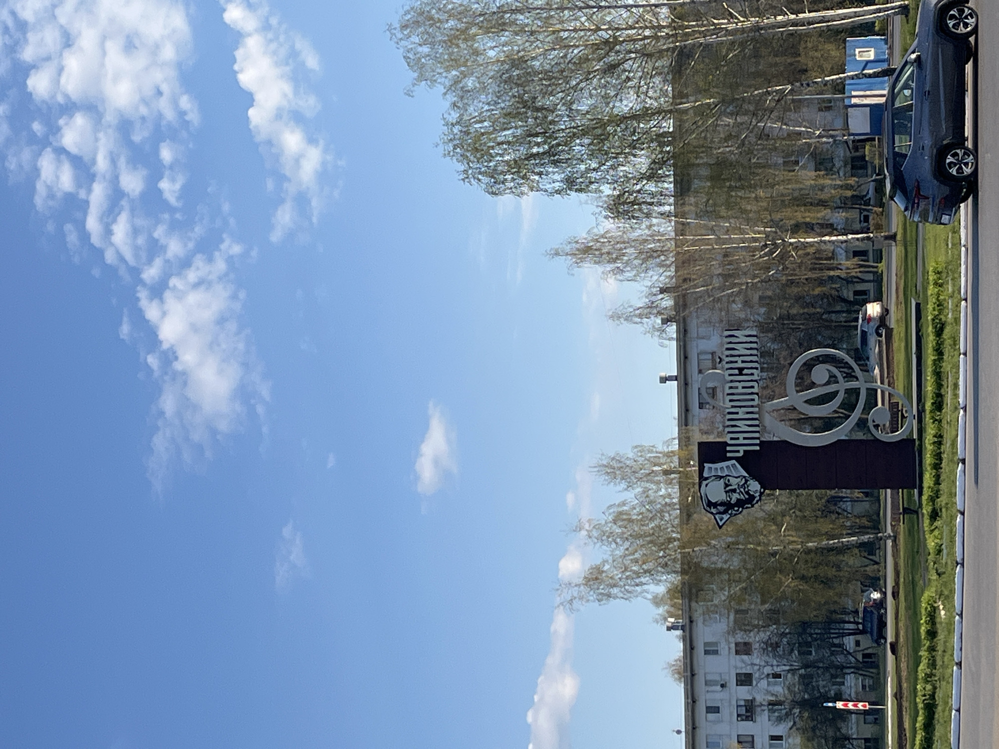 The image depicts the entrance sign to the city of Chaikovsky, Russia. The sign features the name "Чайковский" in Cyrillic script above a stylized musical treble clef and a portrait of the composer Pyotr Ilyich Tchaikovsky. Behind the sign are several multi-story apartment buildings and sparse trees. A blue car is parked on the side of the road. The sky is blue with scattered clouds. The photograph appears to be taken during the daytime. The area around the sign is grassy.