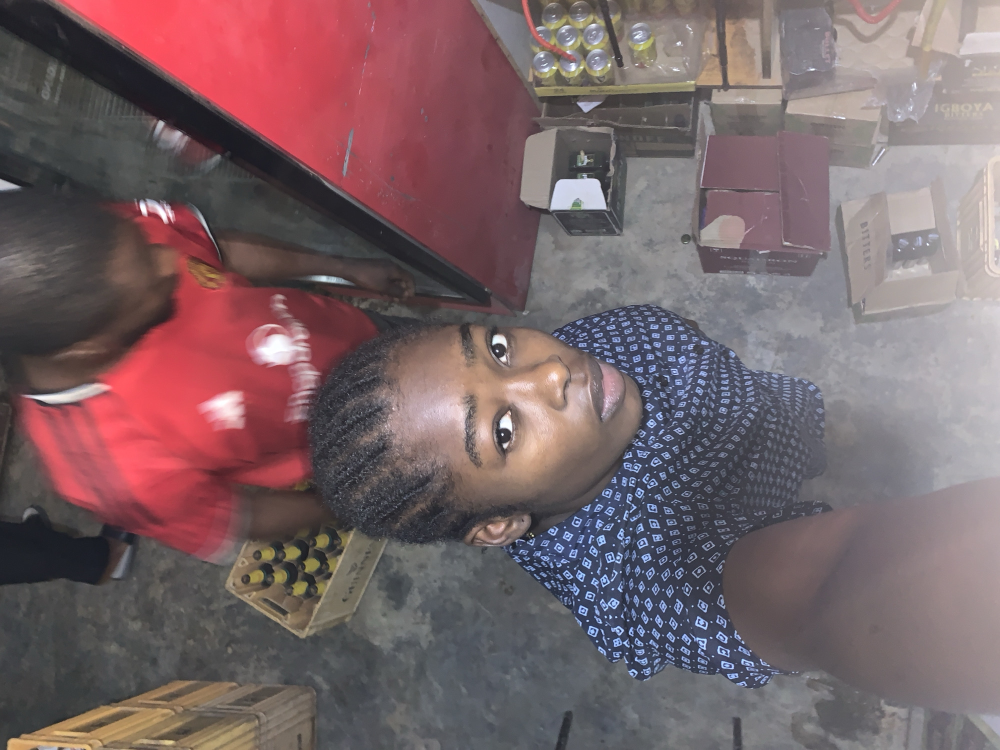 The photo depicts an indoor setting with a young person in the foreground wearing a dark patterned blouse. Adjacent to them, an individual in a red shirt with a white logo appears to be bending over. The immediate environment includes wooden crates, one containing bottles with yellow caps, and various cardboard boxes, some labeled, indicating possible inventory or storage. The overall scene suggests a cluttered or functional space, potentially a small shop or storage area. The city and stadium, New Stadium, Nigeria, are mentioned without being visible or directly discernible from the photo.